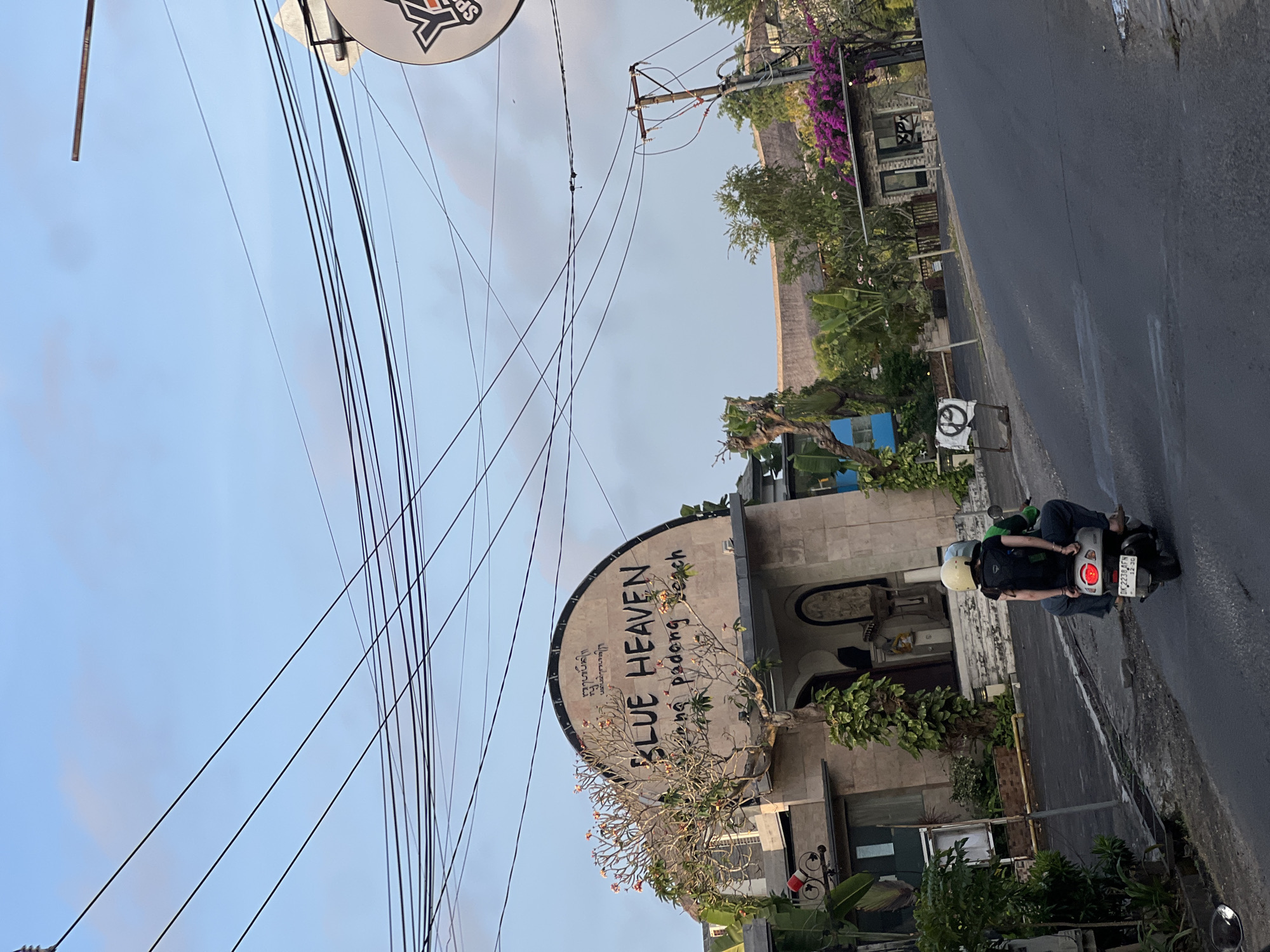 In Kuta Selatan, Indonesia, a street scene unfolds under a hazy sky. A paved road curves to the right, with a slight incline. On the left, a light-colored building with an arched facade prominently displays the text "BLUE HEAVEN" in black lettering, followed by smaller script that reads "nana padang beach". A tree with sparse foliage and small pink flowers stands before the building. Two people, one wearing a yellow helmet and the other a green helmet, ride a scooter away from the camera. The scooter's rear brake light is illuminated. The foreground of the road shows some damp patches. On the right side of the road, a small guardhouse or kiosk with a tiled roof is visible, adorned with vibrant purple flowers. Beyond the kiosk, lush greenery and trees are abundant. Overhead, a complex network of power lines crisscrosses the sky.