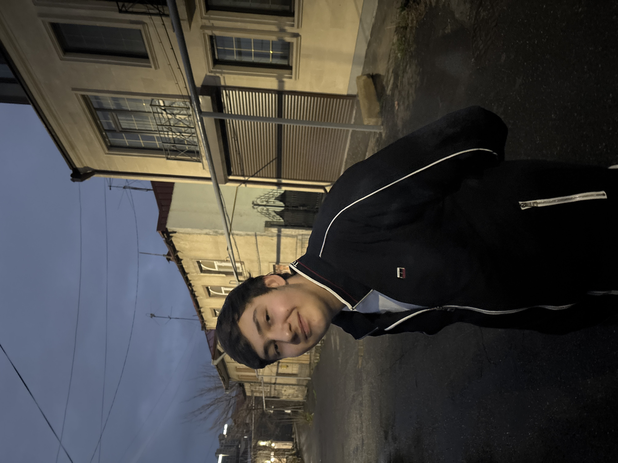 A young man is standing on a street in Yerevan, Armenia. He is wearing a black jacket with white stripes. The street is paved and there are buildings on both sides. The sky is dark.