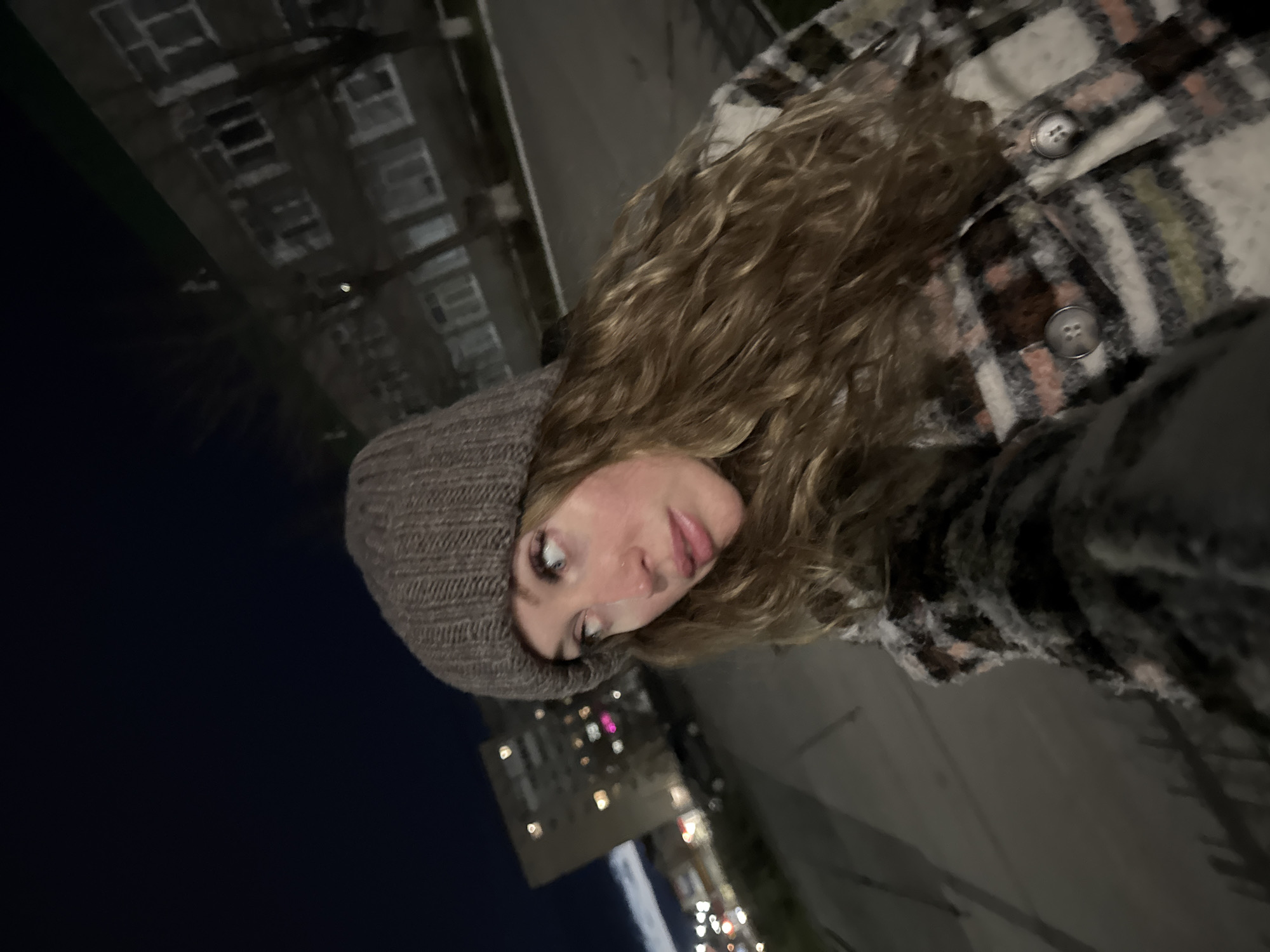 The media file is a nighttime selfie featuring a young woman in an urban setting in Chaikovsky, Russia. The woman is positioned centrally, with her head tilted to her left, looking up and slightly away from the camera. She has long, wavy, light brown hair and is wearing a dark, knitted beanie and a thick plaid jacket or "shacket" with visible buttons. The jacket features a pattern with various shades, including white, black, brown, and hints of pink.
The background is dark, indicating night. To her left, the blurry facade of a building with windows is discernible against the dark sky. To her right and below her head, numerous out-of-focus lights suggest illuminated buildings, streetlights, and possibly car lights in the distance, creating a bokeh effect. The overall image is tilted, giving a dynamic perspective to the scene. The woman's expression is neutral, and her warm attire suggests cool or cold weather. There is no visible text in the image.