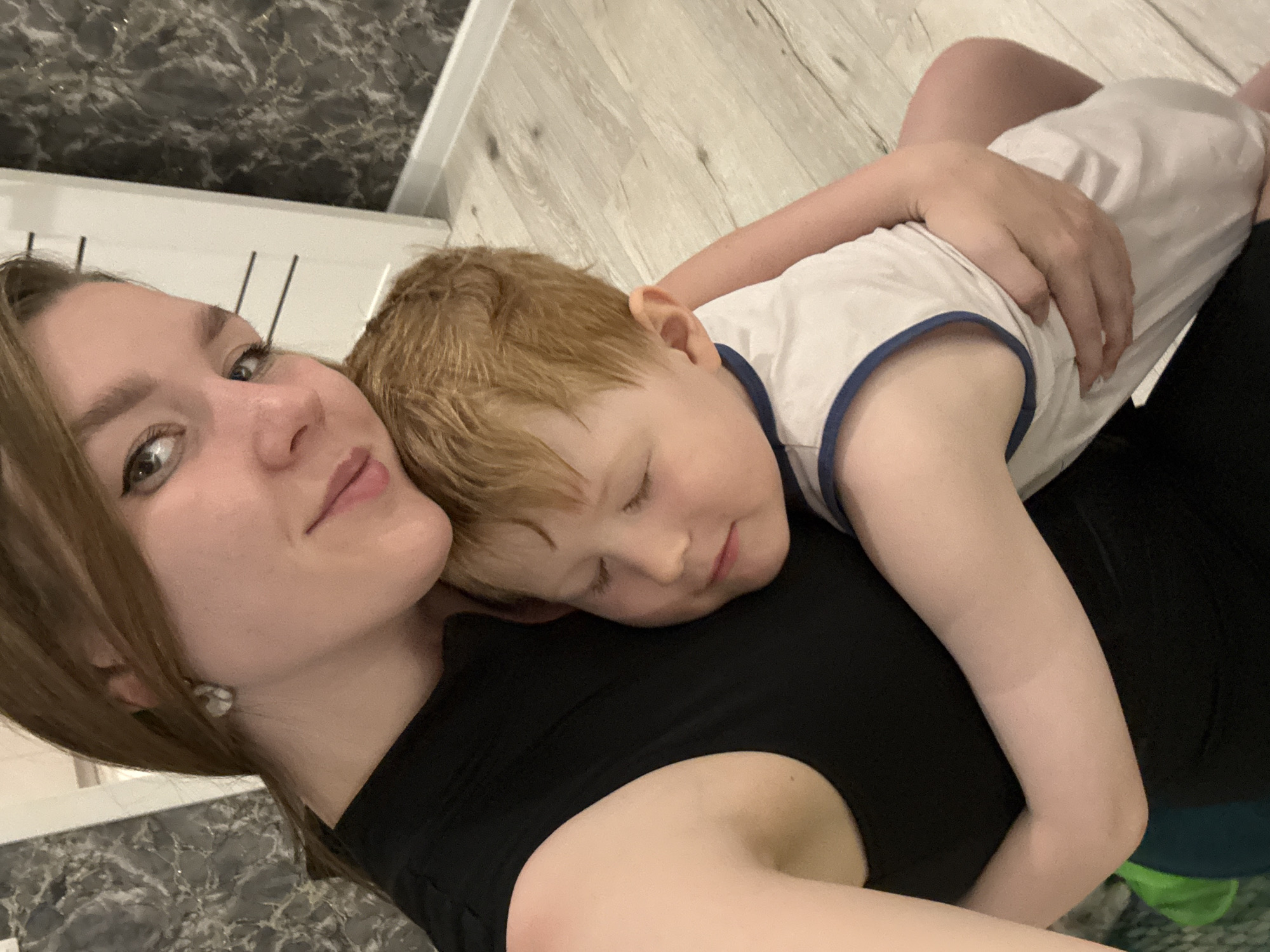 A woman and a young child are lying down together. The woman is on her back, wearing a black top. The child, with red hair, is lying on top of her, seemingly asleep. The child is wearing a white tank top with blue trim. The woman's arm is around the child. They are positioned on a floor with light-colored wood-like panels. The background includes a wall with a dark marble pattern and a white element that appears to be part of a door frame or furniture. The image does not provide information to determine if the location is Izhevsk, Russia.