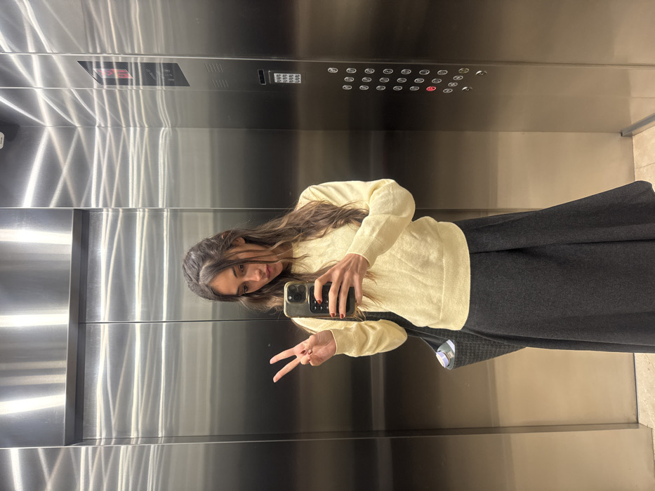 A woman takes a selfie in an elevator. She wears a pale yellow sweater and a long gray skirt. Her long brown hair is styled with a headband. She holds her phone in her right hand and makes a peace sign with her left. The elevator is modern with a mirrored interior and a panel of buttons. The photo is taken at an angle.