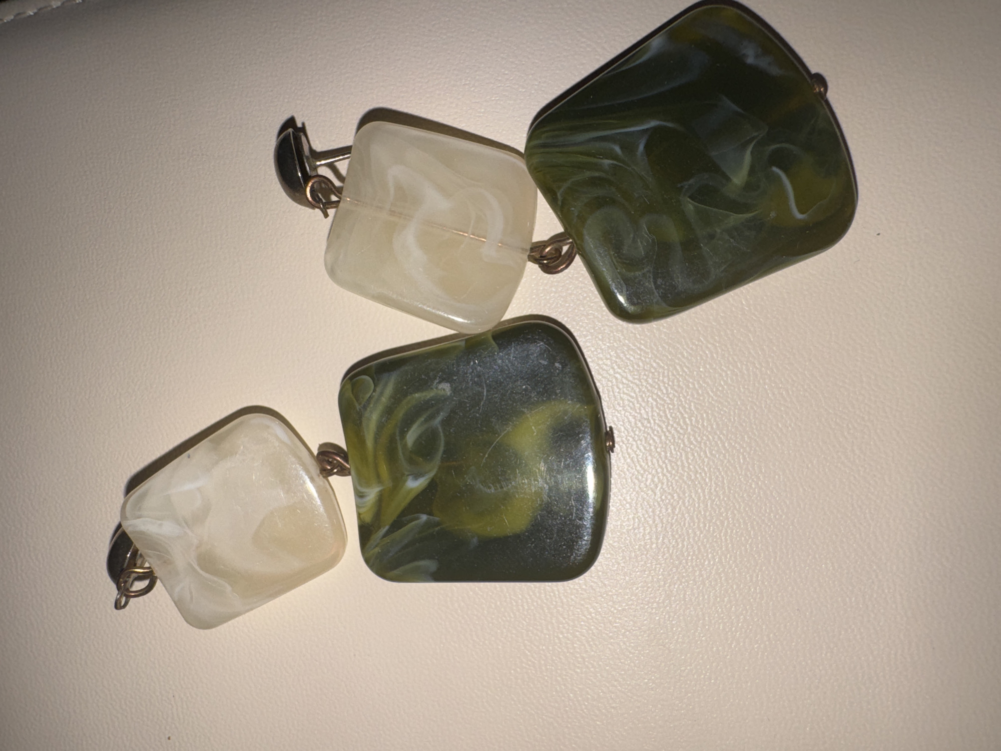 A still life image displays a section of a bracelet featuring four large, square, translucent beads connected by metal loops. Two of the beads are an off-white color with swirling milky patterns, while the other two are a dark green with marbled white and lighter green swirls. The beads are laid out diagonally on a textured, light-colored surface, possibly a leather or faux leather material. The lighting casts shadows, suggesting a light source from the top left. There is no indication of people, activities, or text in the image. The location context, "Яромінскі сельскі Савет, Belarus," is external information and not discernible from the visual content of the image itself.