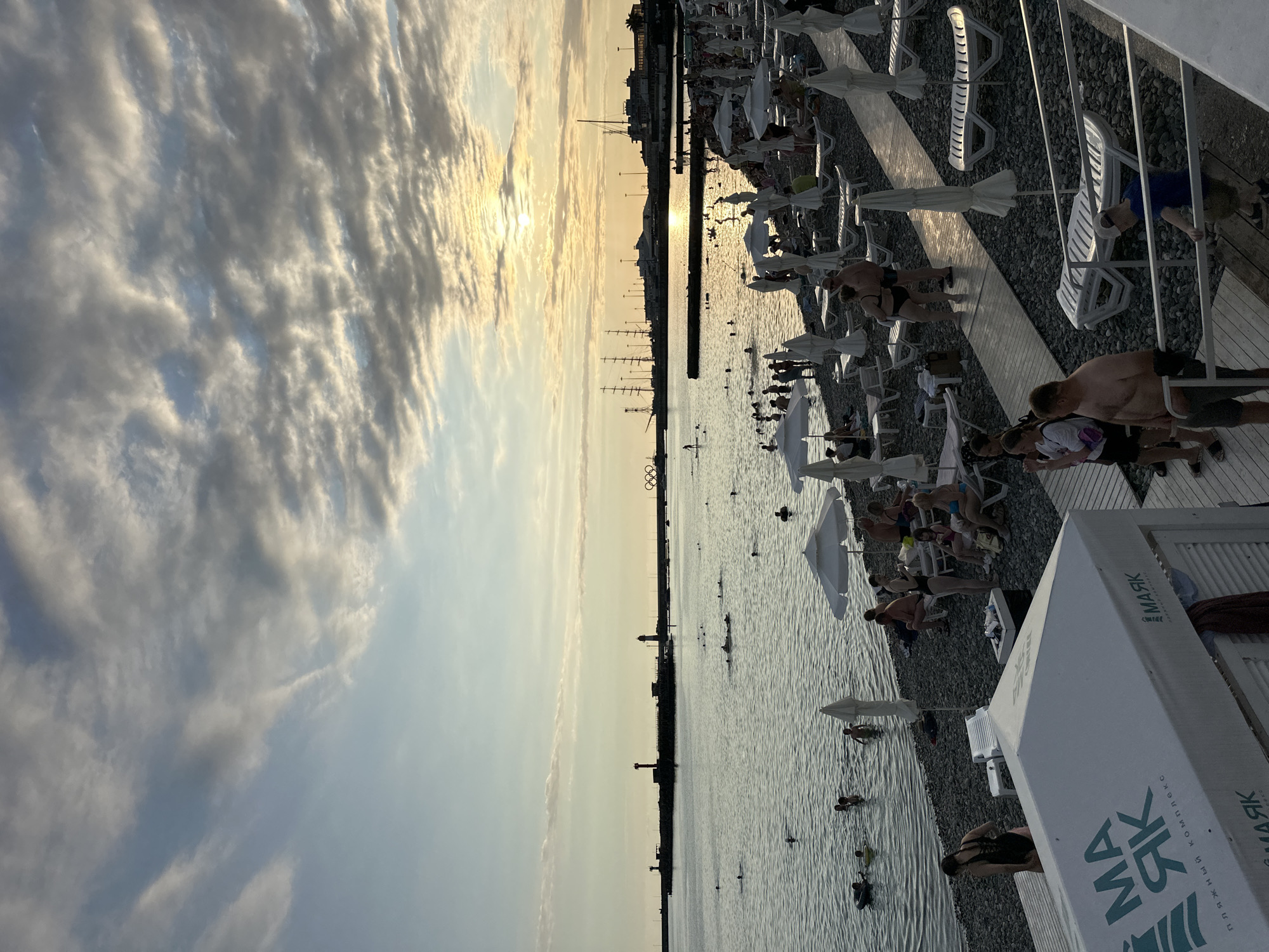 The image depicts a crowded beach in Sochi, Russia, during a sunset. The sun is low on the horizon, casting a warm glow and creating a dramatic sky with scattered clouds. The sea is calm, with numerous people swimming and wading. Beach chairs and umbrellas are set up on the pebbled shore, occupied by sunbathers. In the background, a harbor with lighthouses and ships is visible, along with the Olympic rings structure. The overall scene captures a lively atmosphere of a popular tourist destination.