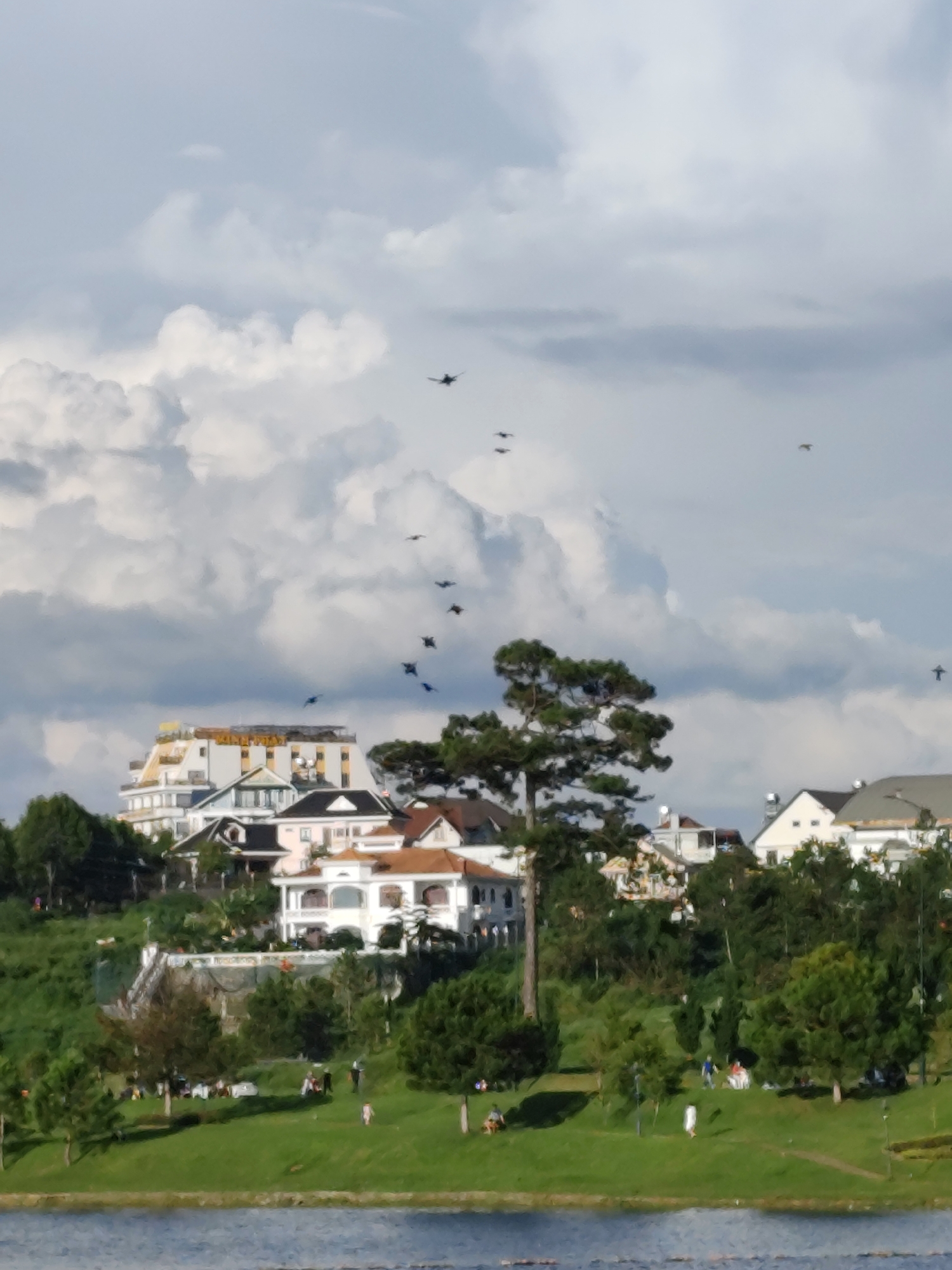 This is a medium shot, slightly angled, full-frame photograph of a cloudy sky above a hillside dotted with buildings and trees, with a body of water in the foreground. The sky is filled with dramatic, billowing clouds in shades of white and gray. Several dark shapes resembling birds or aircraft are scattered across the sky, with some appearing in clusters and others isolated.
The hillside is densely populated with a mix of architectural styles, including a prominent, multi-story white building with yellow accents and balconies at the top, possibly a hotel or large residence. Below this are several smaller houses with pitched roofs in varying colors, including white, pink, and brown. The buildings are nestled amongst lush green trees, with a large, mature pine tree prominently featured on the right side of the frame.
The lower portion of the image shows a gently sloping, grassy park area leading down to a calm, blue body of water, likely Xuân Hương Lake in Đà Lạt, Vietnam. Several small figures, appearing to be people, are scattered across the grassy area. Some are standing, some are sitting, and a few appear to be walking. There are also some picnic blankets visible. The overall impression is a serene, natural landscape with human activity integrated into it, under a dynamic sky.