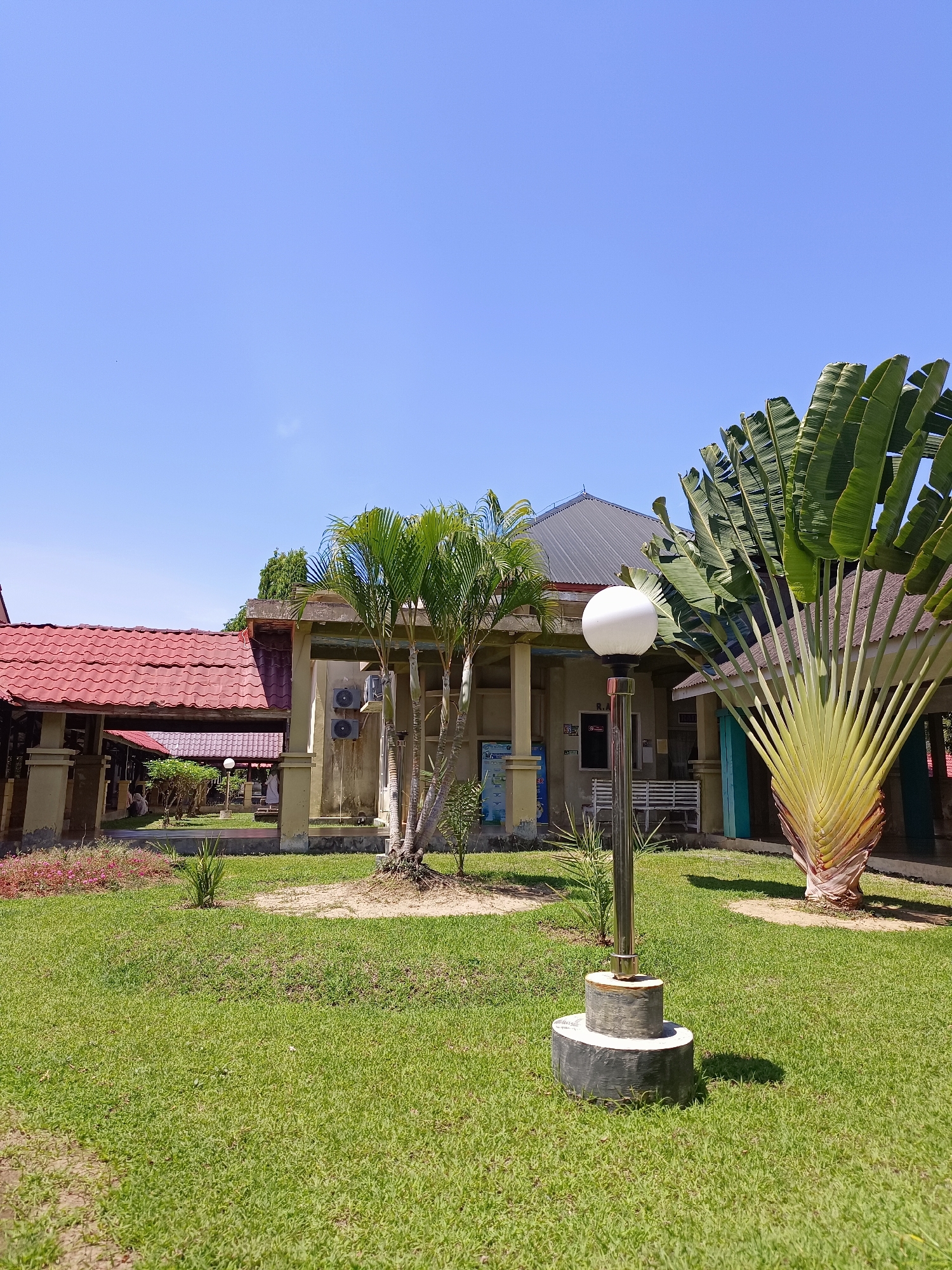 The image depicts a building with a red tiled roof and beige walls under a clear blue sky. In the foreground, a well-maintained green lawn surrounds a modern outdoor lamp post with a white spherical light. To the right of the lamp post, a large fan palm tree with distinctive, paddle-shaped leaves stands prominently. Behind the palm tree and lamp post, part of the building's facade is visible, featuring windows, air conditioning units, and a walkway with pillars. Further into the background, more buildings with red roofs are visible, suggesting a complex or campus-like setting. The environment appears to be warm and sunny, characteristic of a tropical climate. The location is identified as Mesjid Reuntoh, Indonesia. There are no people visible in the immediate foreground or engaging in any specific actions, though faint figures may be discerned in the shaded areas of the building's walkway in the background. The scene is tranquil and architectural, with a strong emphasis on natural elements like the palm tree and the lawn.