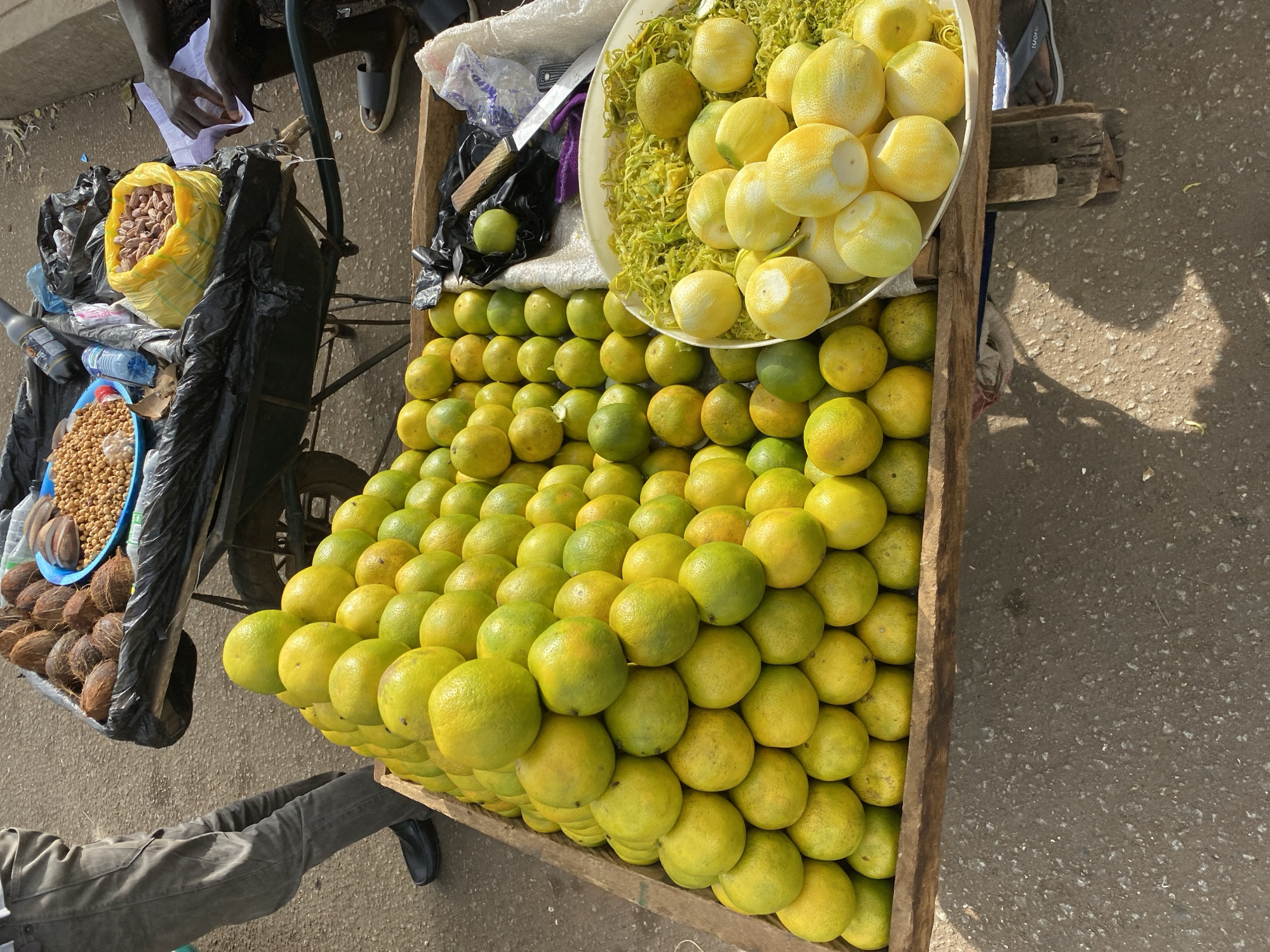 A street vendor in Bukuru, Nigeria, displays a large wooden tray filled with a pyramid-like stack of green-yellow citrus fruits, topped with a plate of peeled citrus fruits and fruit rinds. A knife lies on top of the fruit next to a plastic bag. The vendor's metal cart also holds a bag of peanuts, a bowl of fried chickpeas, and a pile of coconuts, all covered in a black plastic bag. The vendor is standing beside the cart. The scene is brightly lit.