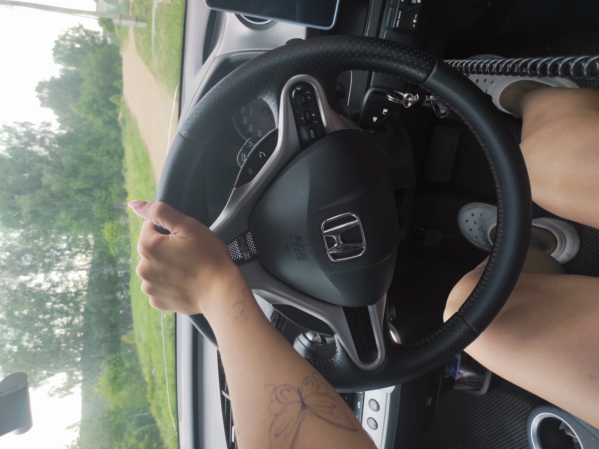 A person is driving a Honda car. The interior of the car is visible, including the steering wheel, dashboard, and center console. The driver's hand is on the steering wheel, and their arm is visible, bearing a tattoo of a butterfly. The driver is wearing sandals. The view through the windshield shows a road and trees. The location is Ольховка, Russia.