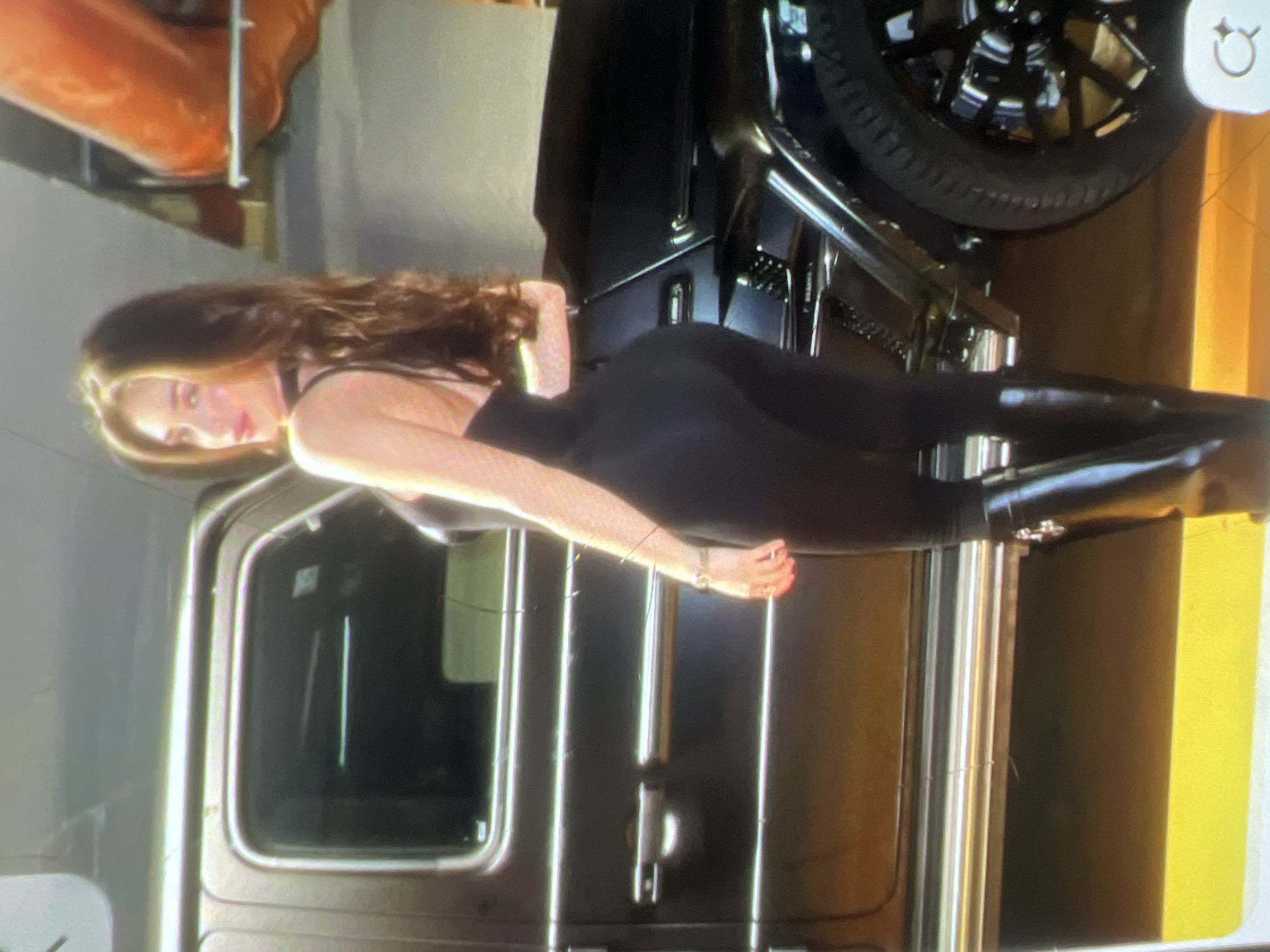 A woman stands next to a black car in an indoor garage. She is wearing a black jumpsuit and black boots. The car is a Mercedes-Benz G-Class. The garage appears to be well-lit with some orange and yellow accents. The woman is looking over her shoulder at the camera.