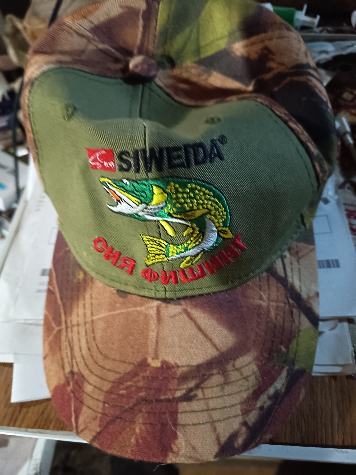 A close-up shows a camouflage baseball cap with a green front panel. Embroidered on the front panel is the word "SIWEIDA" in black capital letters above an image of a fish. Below the fish, there's red Cyrillic text. The cap's brim and the top part have a camouflage pattern of brown, green, and tan blotches. The cap sits atop a stack of papers on a wooden surface. The location is unknown.