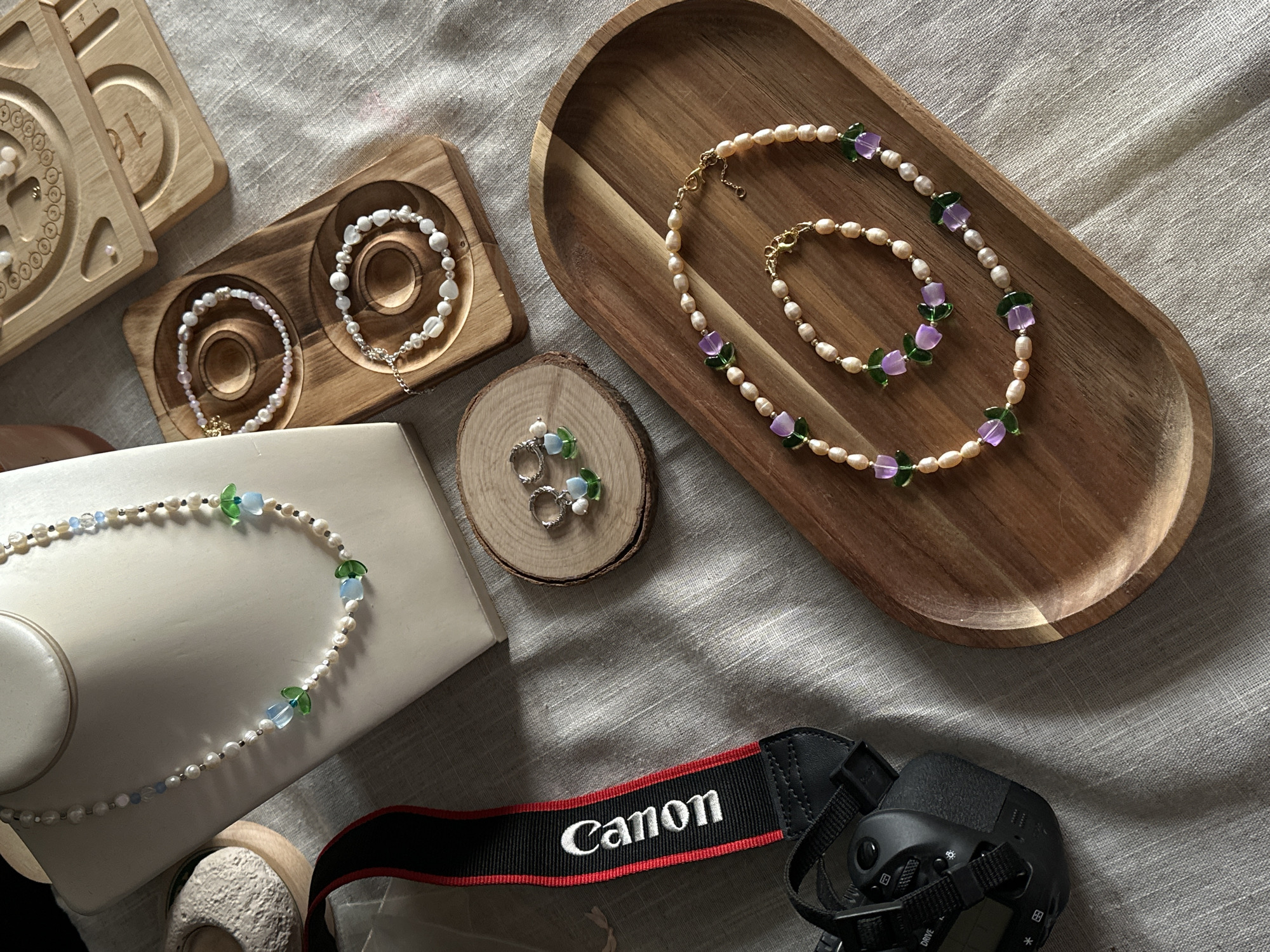 This image captures a flat lay arrangement of various jewelry pieces and a camera, set against a light-colored, textured fabric background, likely indoors in Šušanj, Montenegro.
In the upper right, an oval wooden tray showcases a necklace and a matching bracelet, both crafted from irregular, off-white pearls. These pieces are adorned with striking purple and green flower-shaped beads, and feature gold-toned clasps.
To the left, a rectangular wooden holder with two circular indents displays two different pearl bracelets. One bracelet consists of small, lustrous pearls, possibly with a faint pink hue, and a small gold charm. The other features larger, creamy-white pearls. Nearby, partially visible wooden organizers with carved details suggest a space for jewelry components or display.
In the lower left quadrant, a white, elevated stand presents another pearl necklace. This necklace combines small white pearls with translucent light blue, clear, and green flower-shaped beads, interspersed with tiny silver-colored beads. Adjacent to this, on a small, rustic wooden slice, two silver-toned earrings with delicate light blue and green beads are displayed.
The lower right corner features a black Canon camera body, with its distinctive black strap, prominently featuring the "Canon" logo in white lettering against a red stripe. The scene is well-lit, with soft shadows suggesting natural light. No individuals are present, indicating a focus on product display or photography.