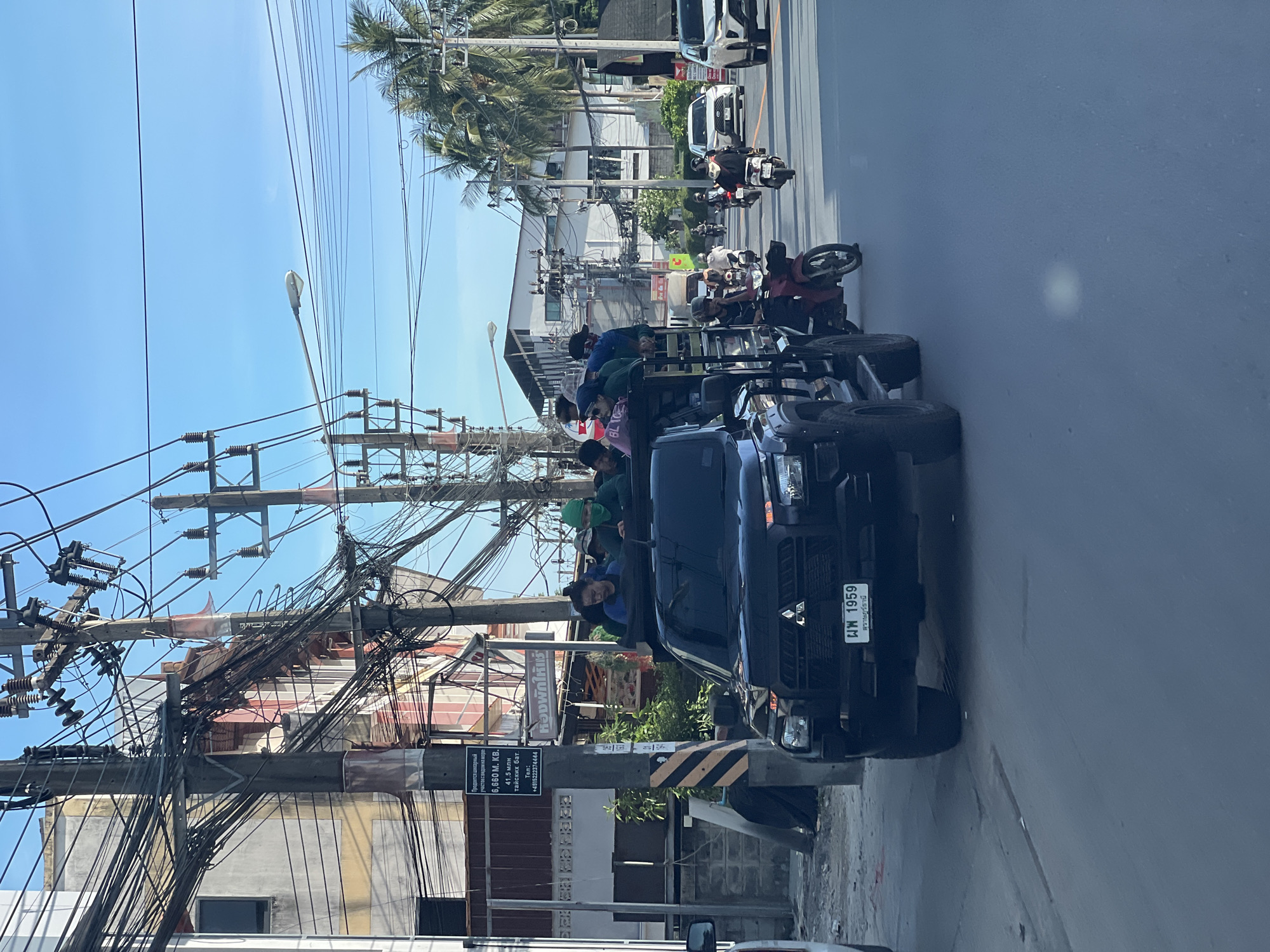 The image captures a scene in what appears to be an urban area, potentially in Southeast Asia, given the presence of Thai script on a sign and the vehicle license plate.
The primary focus is a dark-colored pickup truck, facing forward in the frame. People are visible standing in the back of the truck. The vehicle’s license plate reads "ผพ 1959 สุราษฎร์ธานี".
Behind the truck, the scene extends down a street with other vehicles, including motorcycles and cars. Palm trees line the road.
The left side of the frame shows a utility pole laden with an extensive network of power lines and cables. A sign with Thai text and a phone number is attached to the pole.