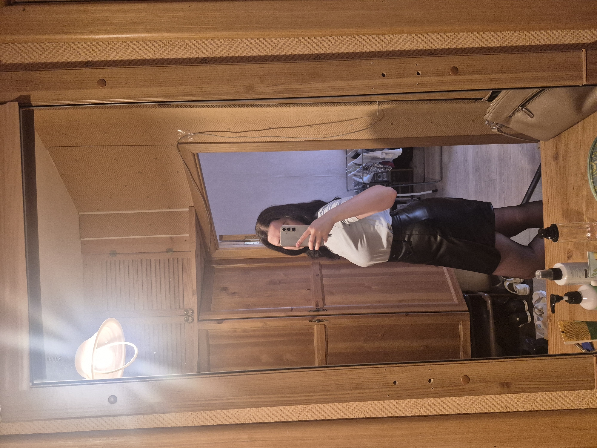 A woman is standing in front of a full-length mirror in what appears to be a bedroom or dressing area. She is wearing a white long-sleeved top and a black leather skirt, and black pantyhose. She is holding a smartphone in her right hand, taking a selfie. The room has wooden paneling on the walls and floor. A light fixture is visible reflecting in the mirror. There is a wooden wardrobe to the left and a doorway to the right, showing a clothes rack with items of clothing. On a surface in front of the mirror, there are several bottles of what appear to be toiletries or beauty products. There is a beige handbag on a wooden surface to the right of the image. The scene does not provide any specific indicators to place its location within the city of Saint Petersburg, Russia.