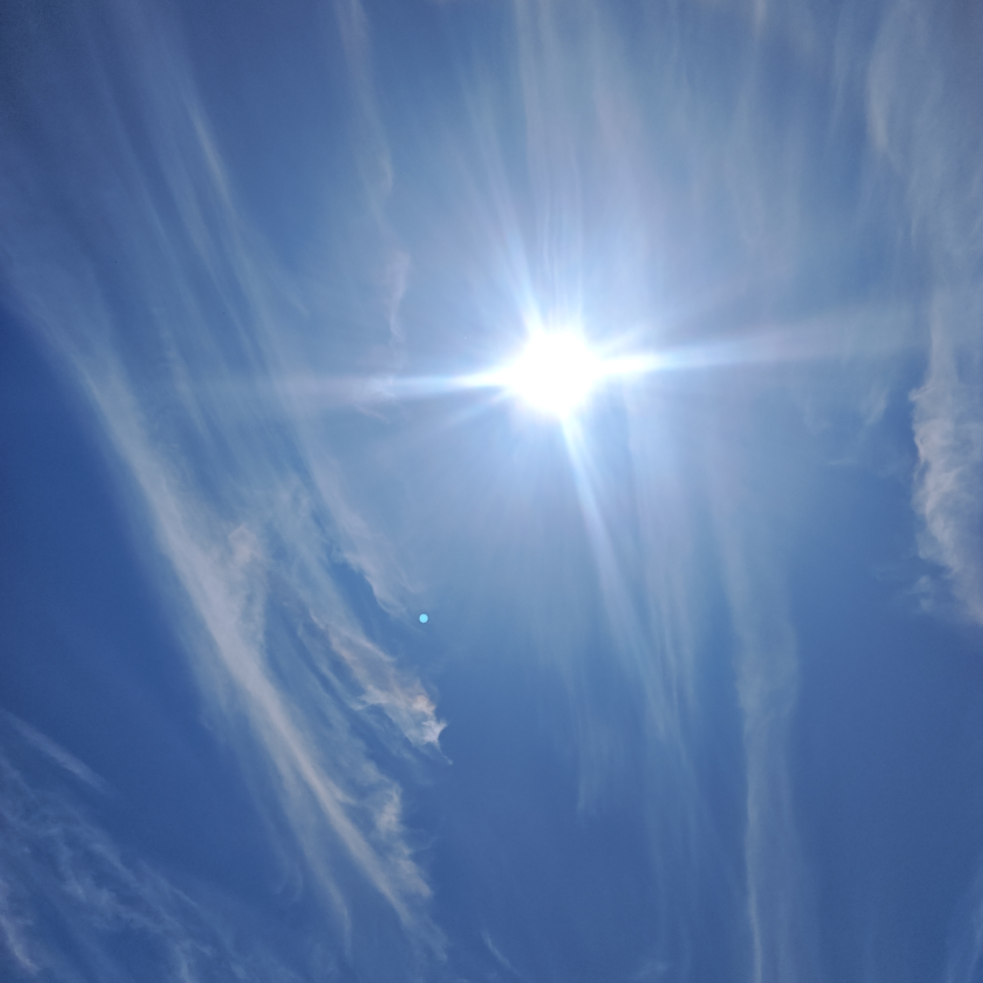 The scene depicts a bright, sunlit sky over Мысовское сельское поселение, Ukraine. The sun, a brilliant white light source, is positioned in the upper half of the frame, emitting a prominent starburst flare with distinct rays. Numerous elongated, thin, white clouds (likely cirrus) streak diagonally across the deep blue sky, appearing most concentrated towards the left and right edges and less dense near the sun's intense glare. A distinct, small cyan-blue circular artifact (lens flare) is visible in the mid-left portion of the frame. The sky transitions from a darker blue in areas away from the sun to a lighter, almost washed-out blue closer to the light source.