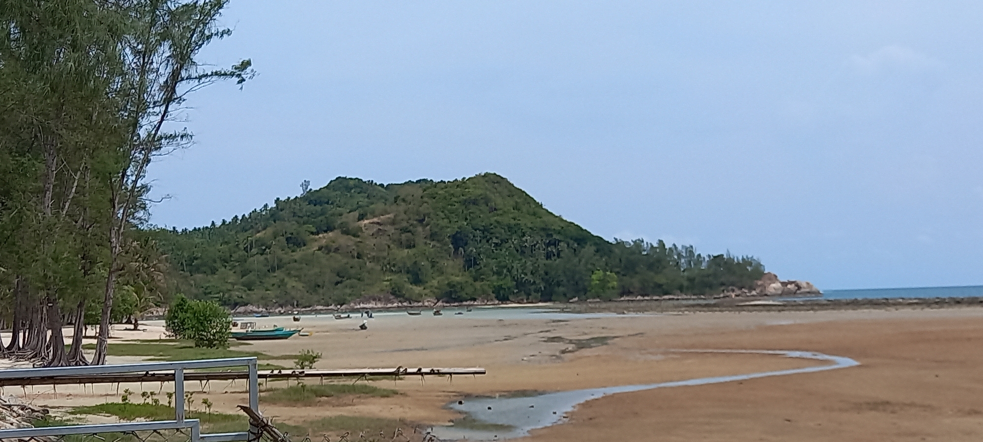 The image shows a coastal scene in Baan Chaloklum, Thailand. A prominent, densely forested hill rises in the background. The foreground features a sandy beach, partly submerged with shallow water creating a stream-like effect across the sand. Several small fishing boats are scattered along the shoreline. Trees line the left edge of the image. A railing is visible in the bottom left corner. The sky is overcast.