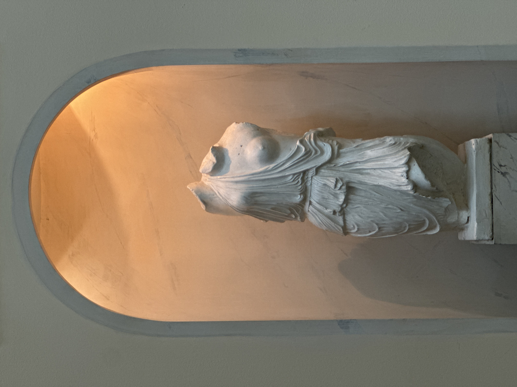 A fragmented white plaster statue torso is nestled within a rectangular niche in a building's wall, with the niche's interior surface painted in a warm, orange hue. The statue appears to be of classical origin, with draped fabric details. The niche is bordered by a gray trim. There is no evidence to support the statue's location in Moscow, Russia.