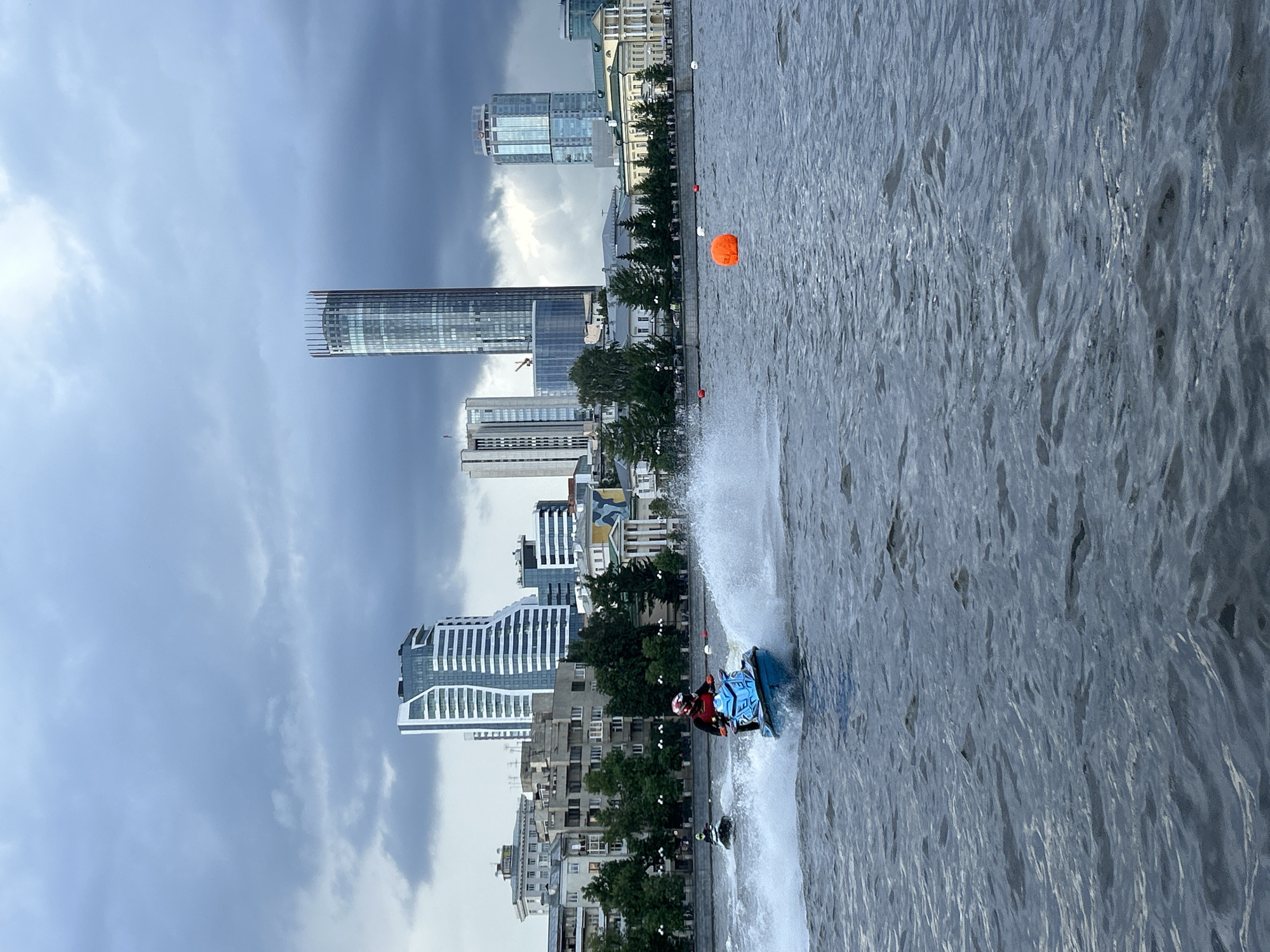 The image captures a dynamic outdoor scene on a large body of water with the city of Yekaterinburg, Russia, in the background under an overcast sky.
In the foreground and midground, two individuals are riding jet skis on choppy, dark blue-grey water. The most prominent jet skier, positioned slightly left of center, wears a red and black suit with a white and red helmet and operates a blue and black jet ski. This jet ski is actively moving, generating a significant white spray of water from its rear. Further back and to the left, a second, smaller jet ski, largely obscured and appearing dark, is also in motion, producing a smaller wake. Several buoys, including a prominent orange spherical buoy in the mid-right and smaller red and white buoys further back, mark a course or area on the water.
The background reveals a diverse urban skyline along the waterfront. Immediately behind the water, a tree-lined embankment separates the river from a mix of architectural styles. On the left, older, multi-story buildings with grey facades and traditional designs are visible. Towards the right, a cream-colored building featuring classical columns and pediments is situated. Dominating the skyline are several modern skyscrapers. The most prominent is the Iset Tower, a tall, cylindrical glass skyscraper with a distinct crown-like top, positioned centrally. To its left, a modern building with a distinctive wavy white facade (likely the Radisson Blu Hotel) stands out. To the right of the Iset Tower, another tall glass skyscraper (likely the Vysotsky Business Center) is visible. The sky above is heavily overcast with dark grey clouds, indicating a cloudy or potentially stormy day.