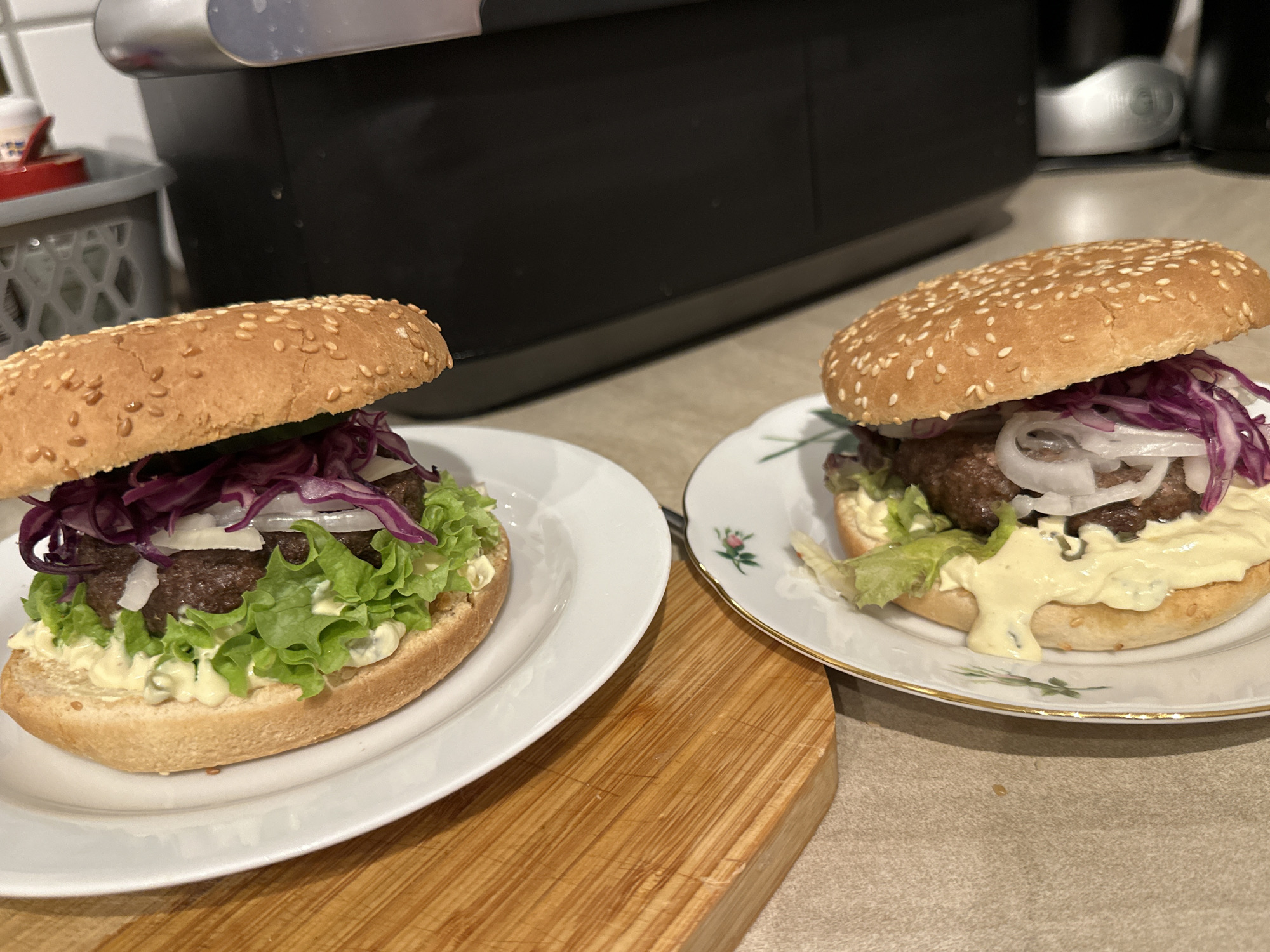 Two homemade burgers are presented on white plates. Both burgers feature sesame seed buns, a beef patty, lettuce, red cabbage, and white onions. The burger on the left is on a solid white plate, while the burger on the right is on a decorative white plate with gold trim and a floral pattern. The burgers are on a countertop, possibly in a kitchen, with a black appliance and some containers visible in the background. A wooden cutting board is placed in front of the left burger. The image does not provide any information to suggest a location in Krefeld, Germany.
