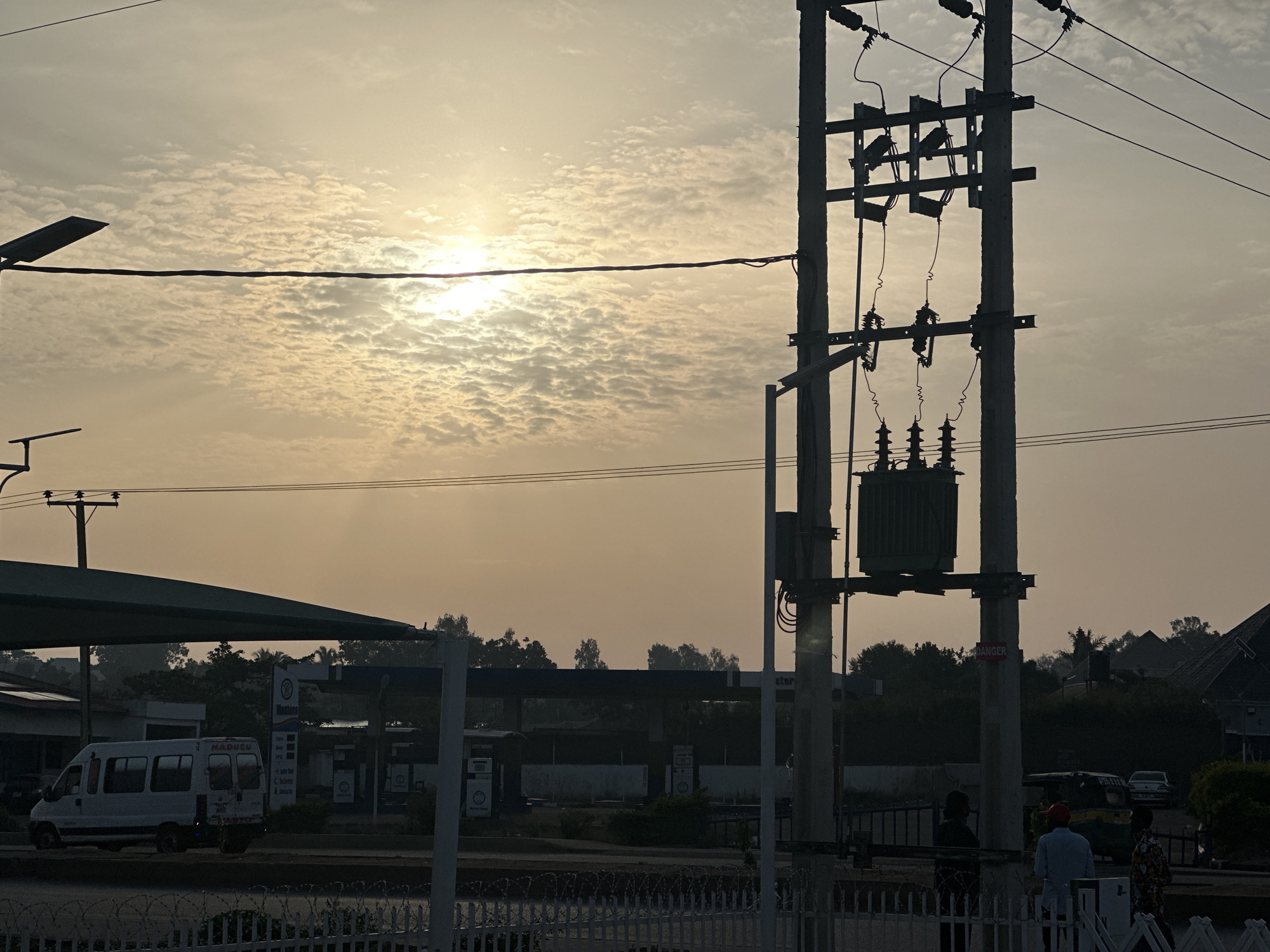 The image shows an outdoor scene in Bukuru, Nigeria, during daylight hours with a bright, hazy sky. A power line structure dominates the right side of the frame. In the background, a gas station is visible, with a white van parked underneath the canopy. Several people are standing near the power line structure. A white picket fence is in the foreground. The overall atmosphere suggests early morning or late afternoon.