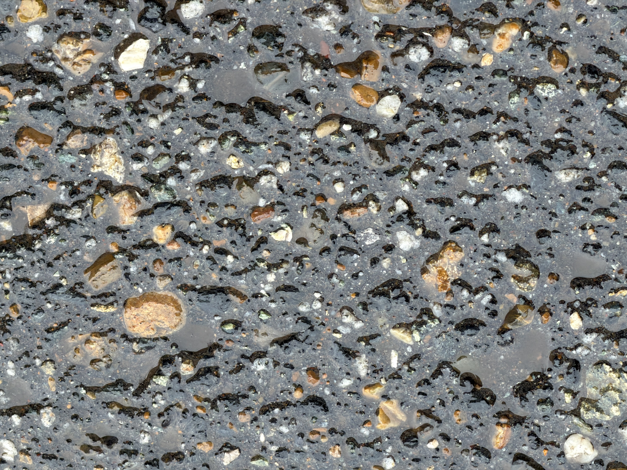 The image shows a close-up of wet asphalt, speckled with small rocks and pebbles. Water puddles collect in the rough surface. The environment is a street. No people or actions are visible in the image. Location within Izhevsk is not discernible from the image.