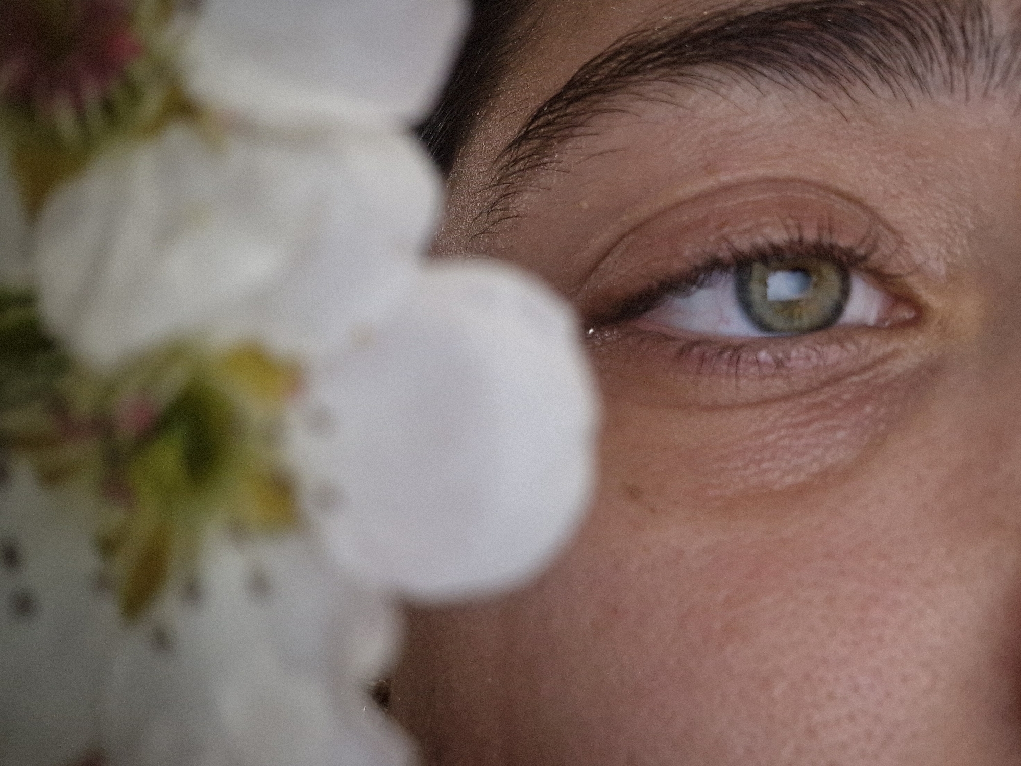 The image shows a close-up of a person's face, with a focus on their right eye. The eye is hazel, with light green and brown hues. The eyebrow is dark and well-defined. The skin around the eye shows some texture and fine lines. Part of the face is obscured by a blurry cluster of white flowers. The location is not identifiable.