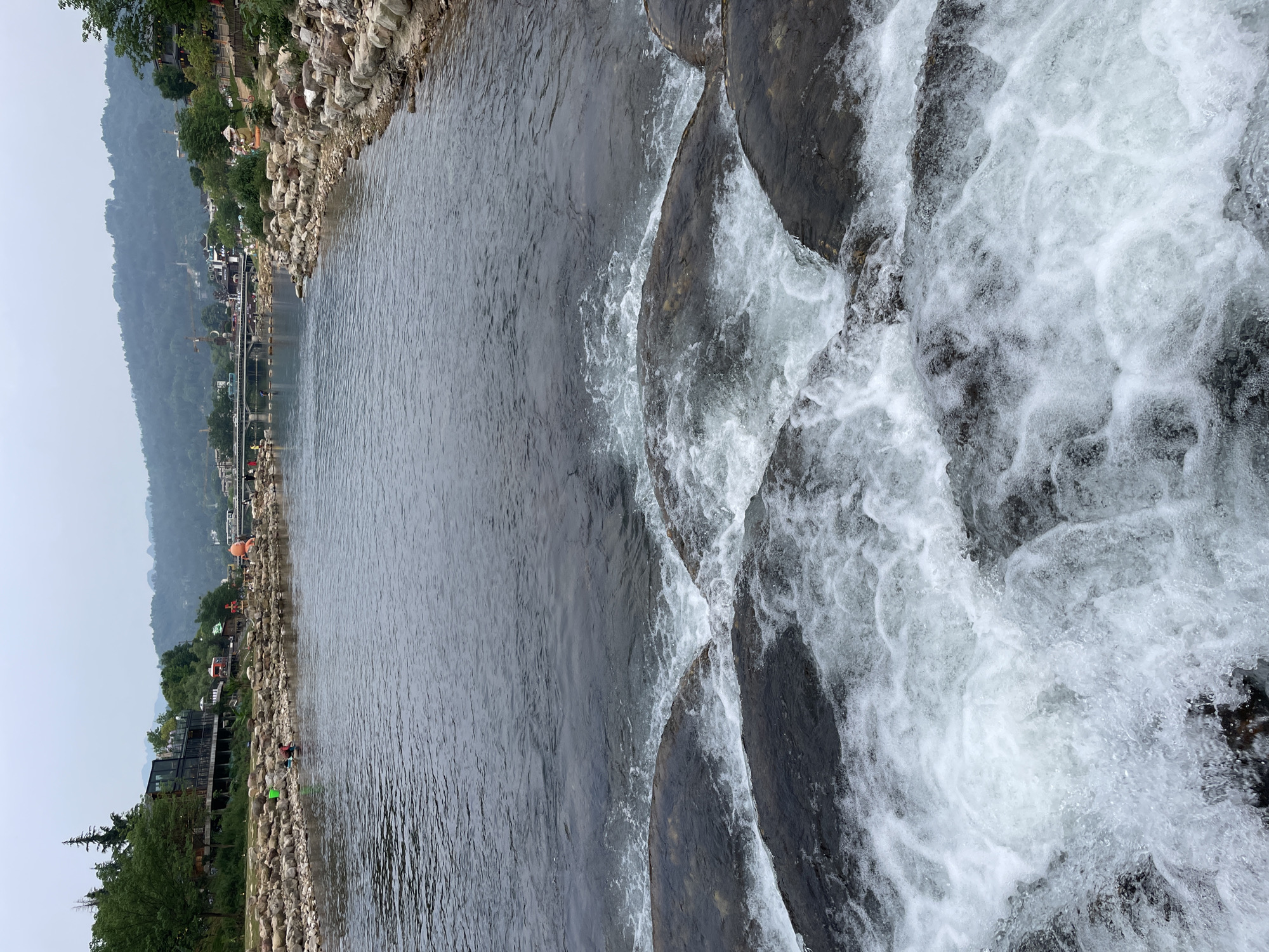 This image shows a water scene at the Suoxi River in Wulingyuan, China. The river flows from the left toward the right, where it cascades over a series of dark rocks, creating white water rapids. The left bank of the river is lined with large, rounded rocks and boulders. Beyond the rocks, there are structures that look like restaurants, and trees lining the river bank. In the distance, a mountain range can be seen beneath a hazy sky. There are some people visible along the river bank, but they are small and indistinct.