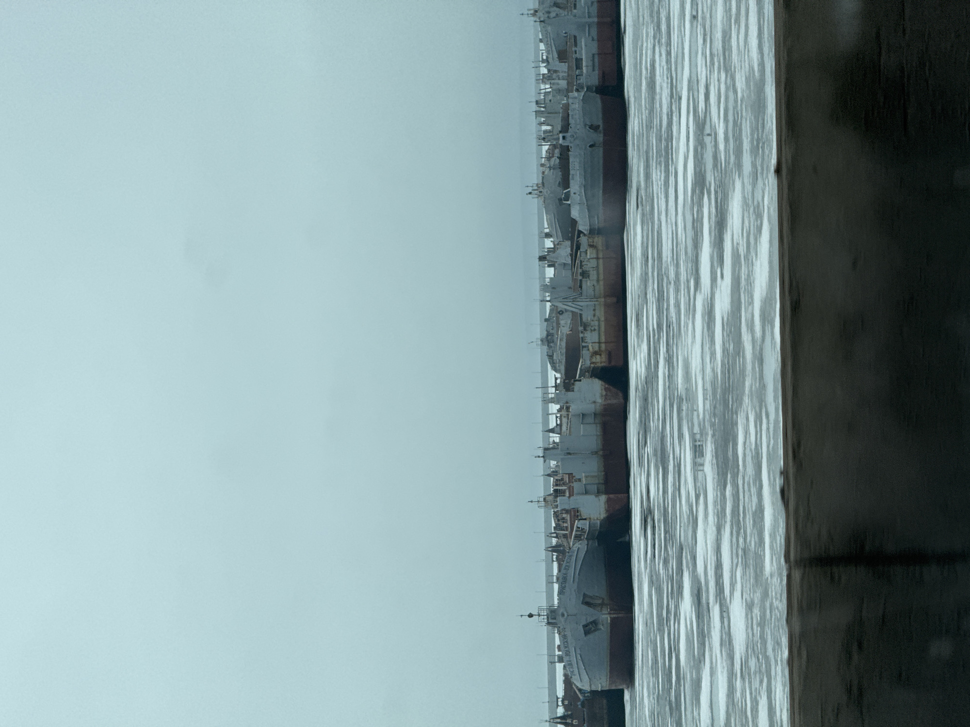 A view from a waterfront in Chaikovsky, Russia, shows a series of large, grey, rusted ships moored side-by-side, extending into the distance. The water surface is choppy, reflecting light. A dark, wooden structure is partially visible in the foreground, obscuring part of the view. The sky is overcast.