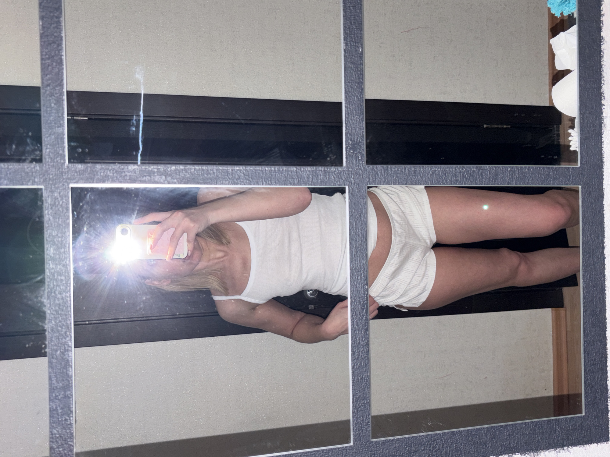 The image is a mirror selfie of a person wearing a white tank top and white shorts. The person is holding a phone with the flash on, obscuring their face. The background consists of a gray wall with a black horizontal stripe. A toilet with a roll of toilet paper is visible in the upper right corner. There is no evidence to indicate the location is the city of Chaikovsky, Russia.