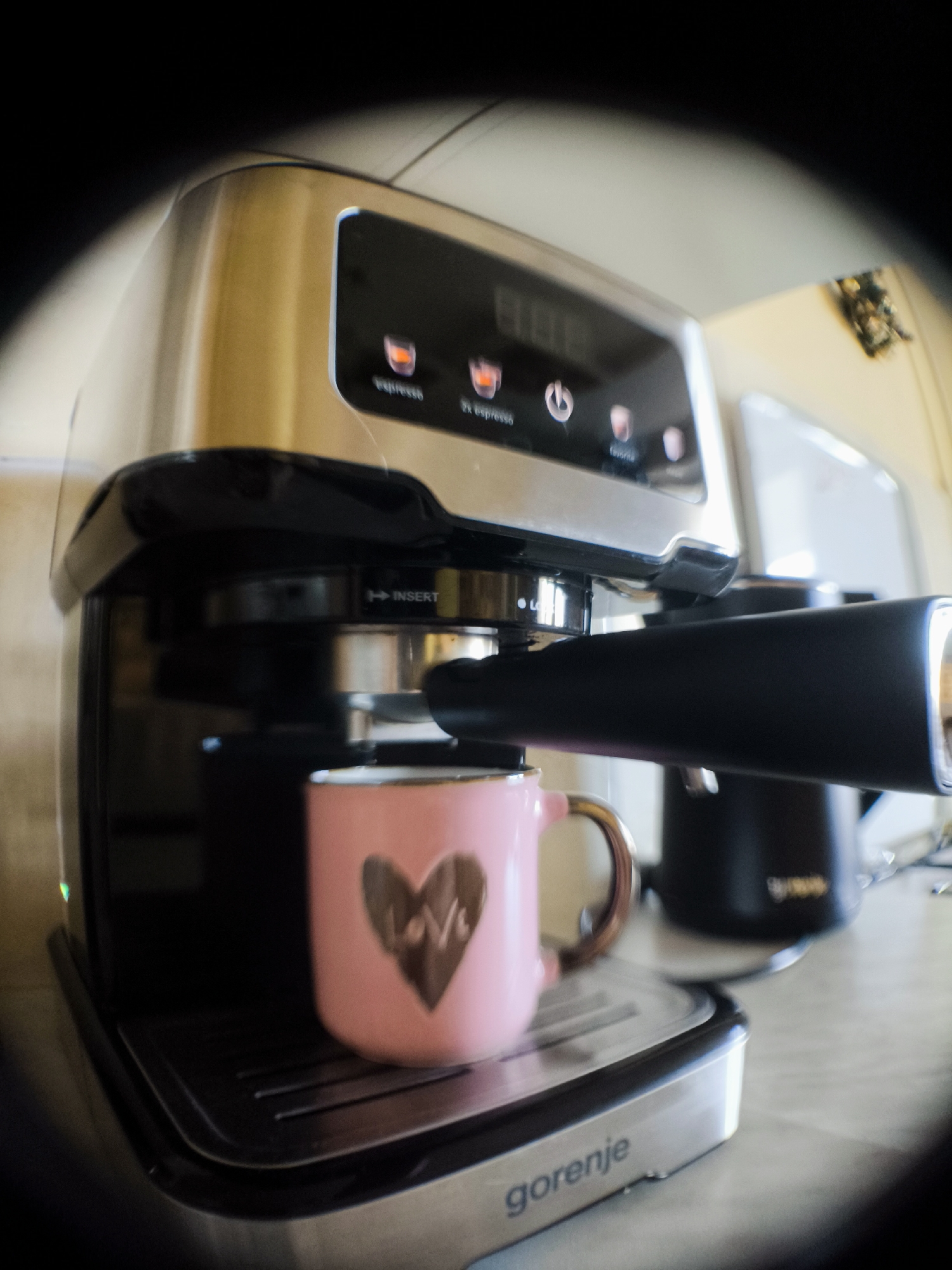A fisheye lens perspective captures a close-up, low-angle view of a Gorenje espresso machine. The machine is primarily silver and black, with a digital display on its top panel showing "888". Below the display are several touch-sensitive buttons with icons representing espresso options, a power button, and a "favorite" setting. The espresso machine has a black portafilter attached, with a silver "INSERT" label visible. A pink ceramic mug with a brown heart design and the word "Love" inside it sits on the drip tray, directly beneath the portafilter, suggesting it is either being filled or has just finished being filled with espresso. To the right of the espresso machine, partially visible, is a black kettle with a silver handle and base, also on the countertop. The background is out of focus, showing a light-colored wall and possibly other kitchen appliances. The overall scene is indoors, likely in a kitchen.