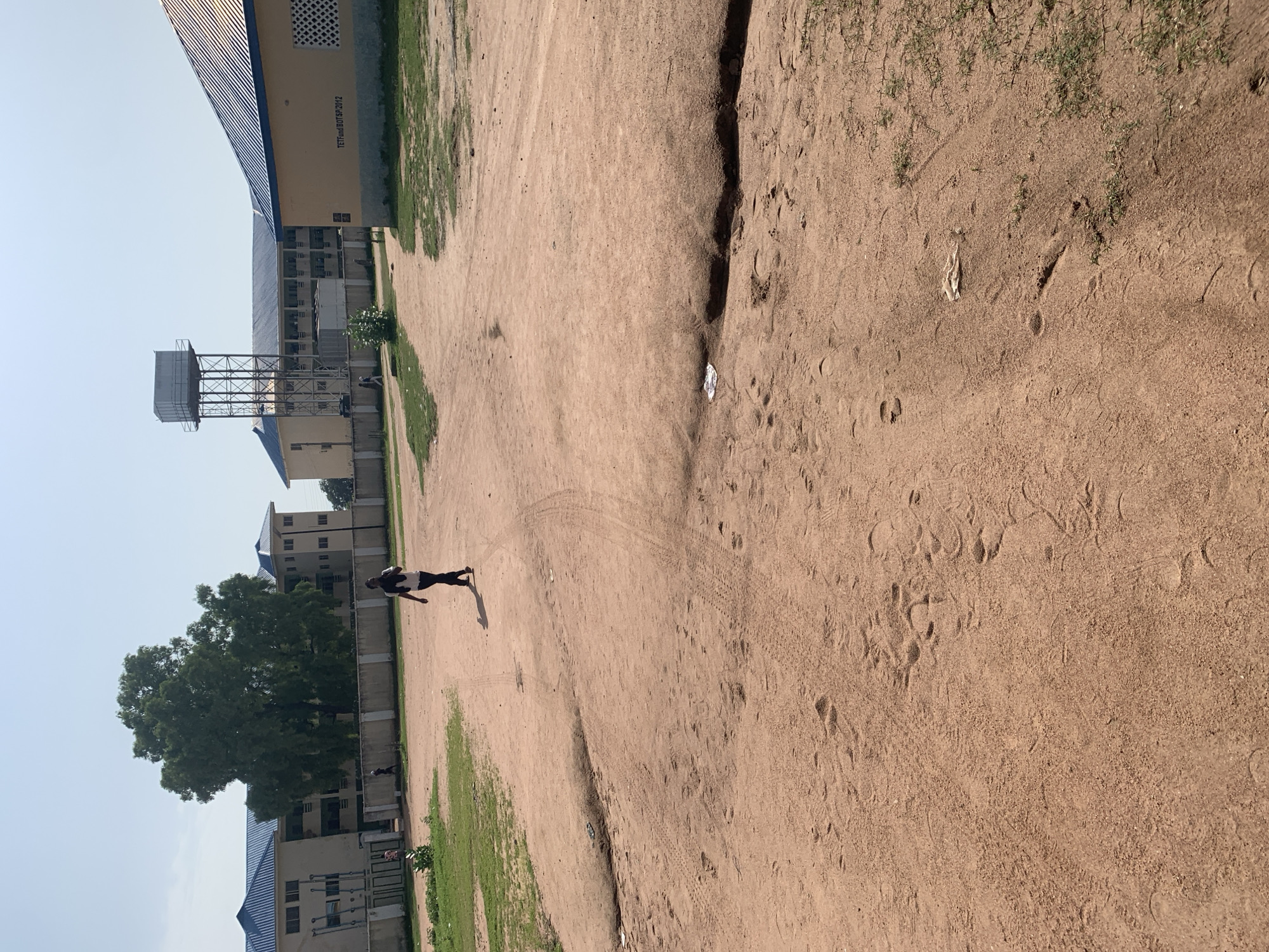 The image depicts an outdoor scene in Jalingo, Nigeria, likely within a university or institutional campus. A person is walking across a dirt and grass area towards the left side of the frame. The background features several buildings with blue roofs and a water tower. The ground is predominantly reddish-brown dirt with patches of dry grass. Tire tracks and footprints are visible on the dirt path. The sky is clear and blue, indicating a sunny day. The buildings appear to be multi-story educational or administrative facilities. One building has text visible on its exterior wall: "TETFUnd BotSP.2012".
