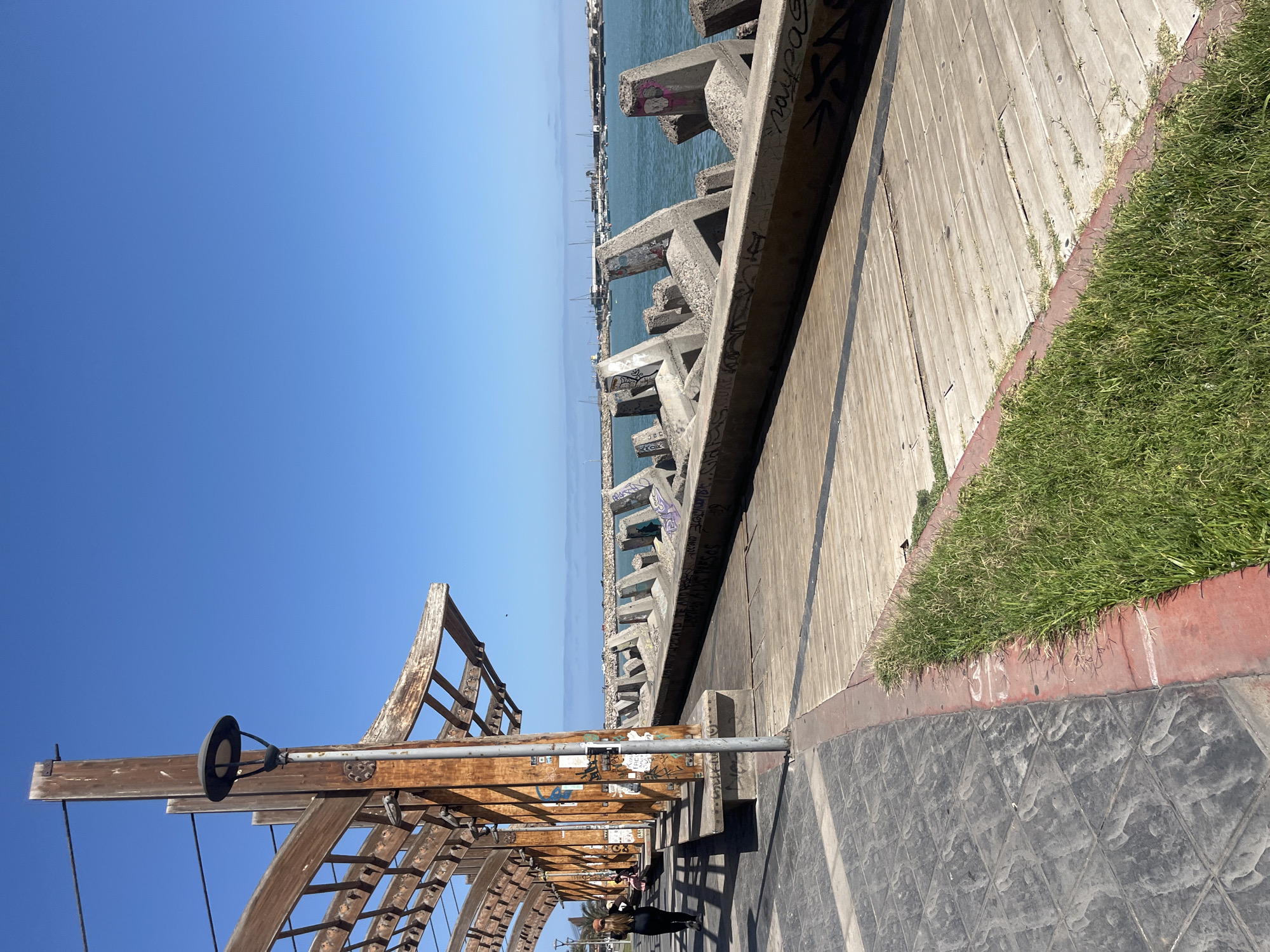 This image depicts a waterfront scene in Arica, Chile. The view is taken from a promenade or walkway lined with wooden structures, possibly part of a decorative archway or pergola. To the right of the walkway is a narrow strip of grass bordered by a red-painted edge, with the walkway surface itself being paved with dark tiles. Beyond the wooden structure, a concrete barrier comprised of interlocking, irregularly shaped blocks runs alongside the water's edge. Graffiti is visible on the concrete blocks. In the background, the ocean stretches towards the horizon under a clear, blue sky. In the lower right of the image, at least one individual can be seen walking along the walkway, and palm trees are visible in the far distance.