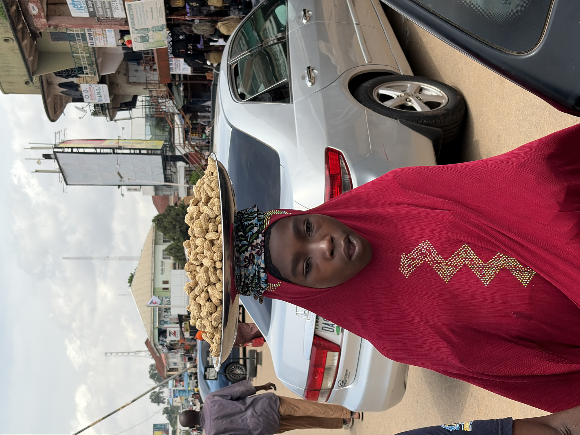 In Jos, Nigeria, a young girl wearing a red hijab and dress, adorned with a zig-zag pattern of gold and white beads on her chest, carries a large metal tray piled high with peanuts on her head. She is walking on a paved street. Behind her, a silver sedan is parked, and further in the background, buildings line the street, with shops and advertisements visible. The sky is overcast. Another person, a man in a gray shirt and brown pants, walks away from the camera in the middle ground. The scene appears to be a typical street market in the city.