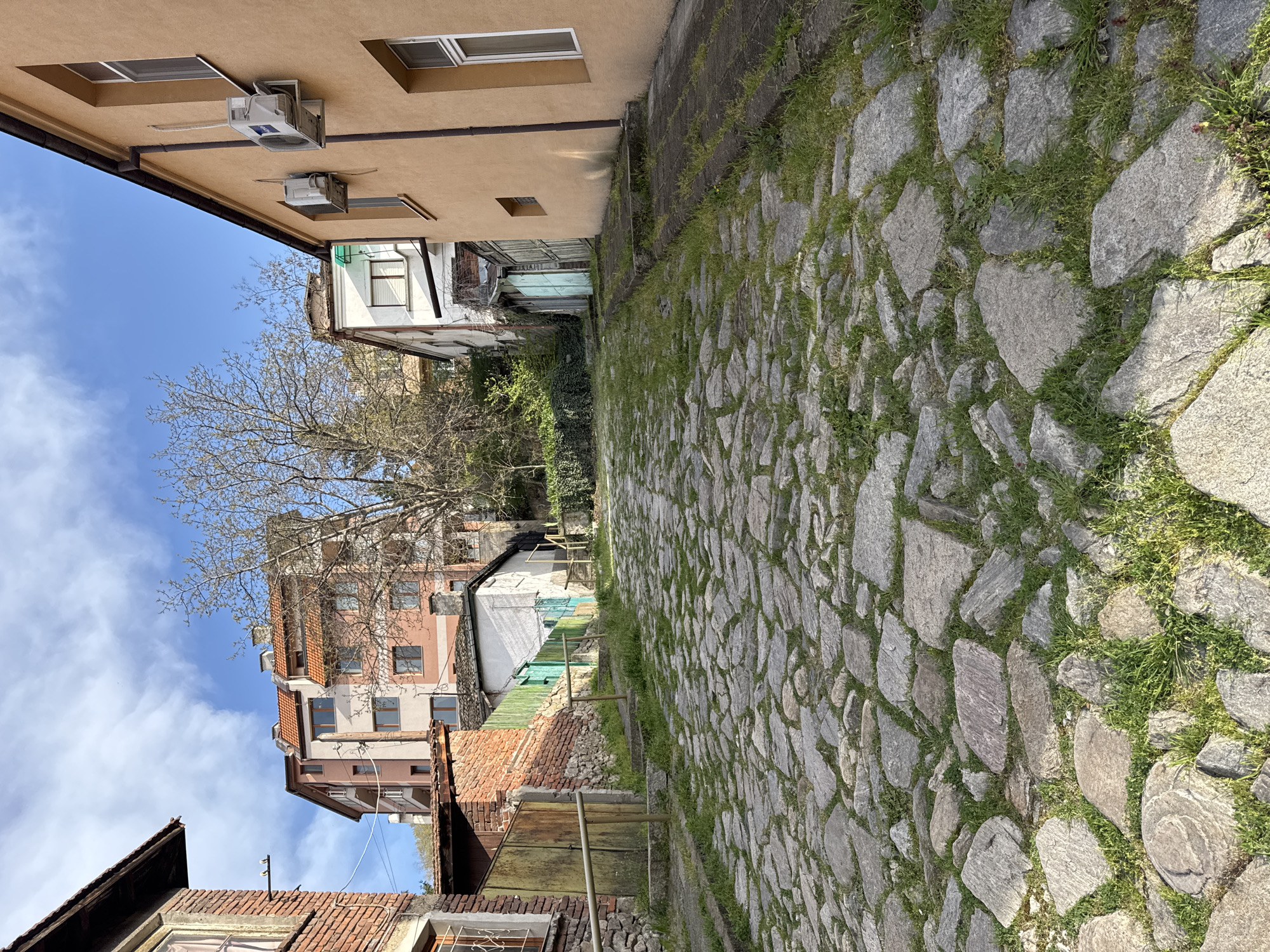 The image shows a narrow cobblestone street in Plovdiv, Bulgaria. The cobblestones are gray and irregularly shaped, with grass growing between them. Buildings line both sides of the street. The building on the left is a light brown color with windows and air conditioning units. Further down the street on the left, the buildings vary in color and design, including a building with pink and white facades. On the right side of the street, there's a concrete wall with grass and vegetation. The sky is blue with some clouds visible above the buildings. A bare tree is visible amid the buildings. The image gives a sense of the old and narrow streets characteristic of Plovdiv's historic areas.