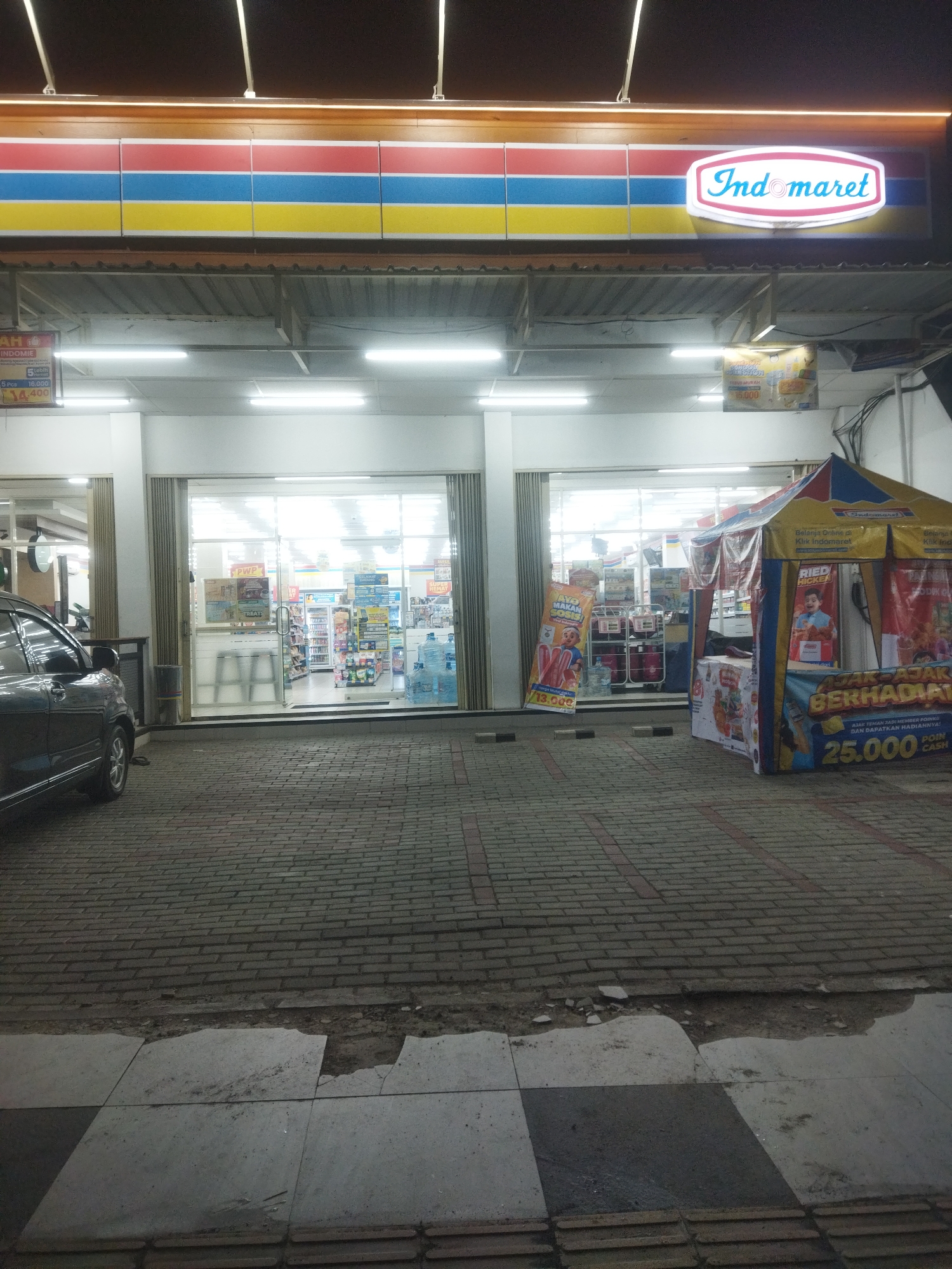 The image shows the facade of an Indomaret convenience store located in Wonogiri, Indonesia. The store has a clean and well-lit interior, visible through large glass doors. In front of the store, a promotional tent is set up, displaying various advertisements and offers. A dark-colored car is parked to the left of the store. The ground in front of the store is paved with bricks. The overall scene is one of a typical evening at a retail establishment.