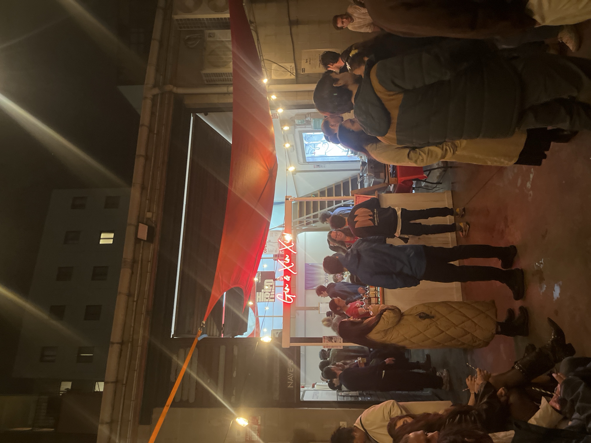 The image shows a nighttime scene at an outdoor bar or event in Barcelona, Spain. The crowd of people is dense. Some people are standing and drinking, while others are seated at tables. Above the entrance is a red awning and a neon sign that reads "Gin & Xin Xin X". The bar or event is located under a large building, and there are other buildings visible in the background. The lighting is artificial, with string lights and spotlights illuminating the area.