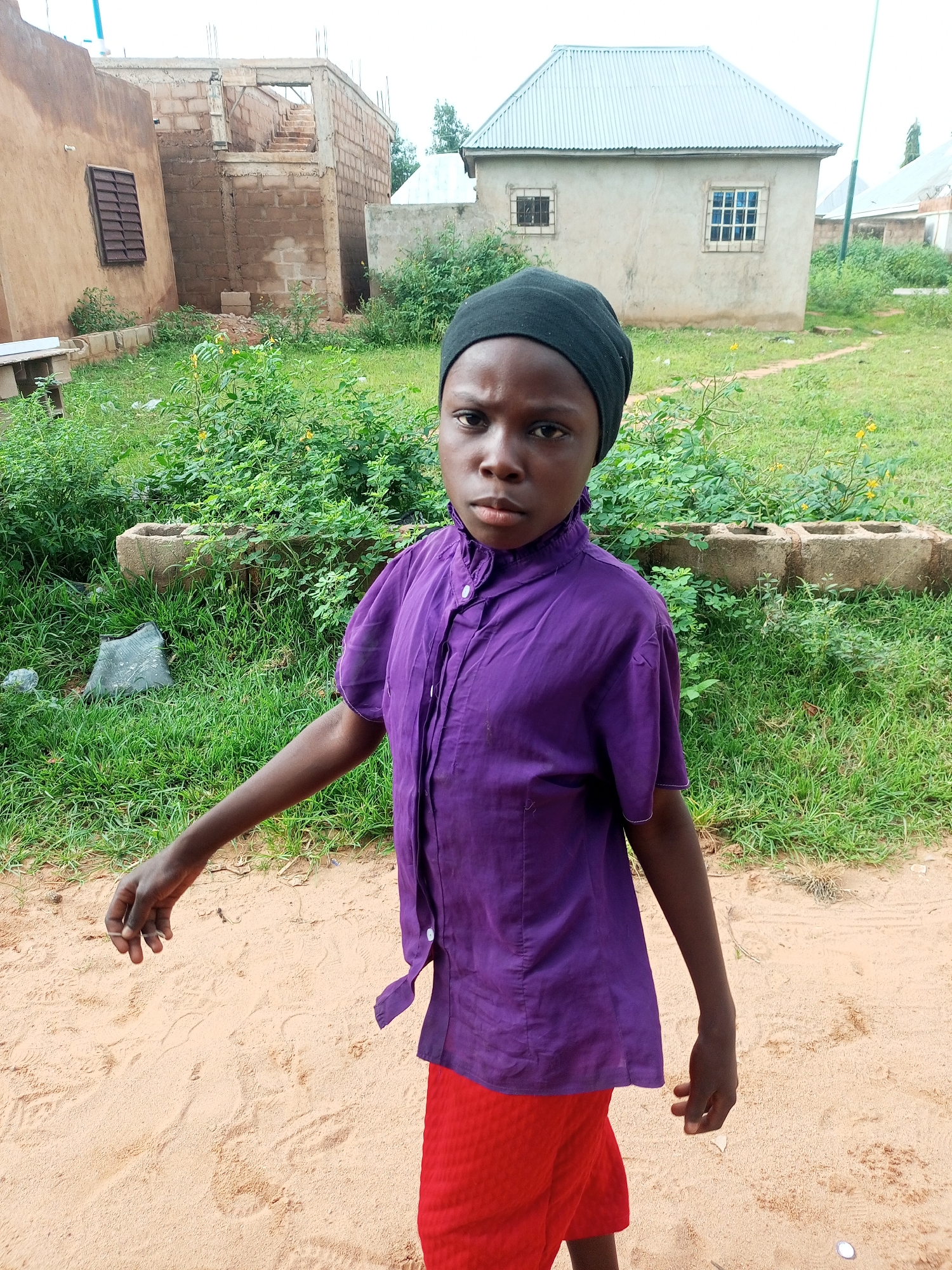 A child wearing a purple shirt and red shorts stands on sandy ground. The child has dark skin and is wearing a black head covering. The child is looking directly at the camera. In the background, there are several buildings under construction, some with brick walls and concrete foundations. There is also a finished building with a corrugated metal roof and windows. Green grass and shrubs are visible throughout the scene. The location is the city of Tempure, Nigeria.