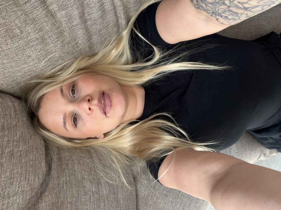 The image presents a selfie-style portrait of a fair-skinned female subject with long blonde hair, reclining in an indoor setting. She is oriented face-up, directly towards the camera. Her eyes are open, and her lips are slightly parted, displaying a light lip color. A small stud earring is visible in her left earlobe (viewer's left). The subject is wearing a plain black short-sleeved top, with a dark colored garment visible beneath the waistline. A complex tattoo featuring floral or mandala-like patterns is present on the inner side of her left forearm (viewer's right). The background is composed entirely of a textured, light brown upholstered fabric, consistent with a couch or similar furniture. No additional environmental details or objects are discernible. The image contains no visual information to determine a specific city location, including Sarapul, Russia.