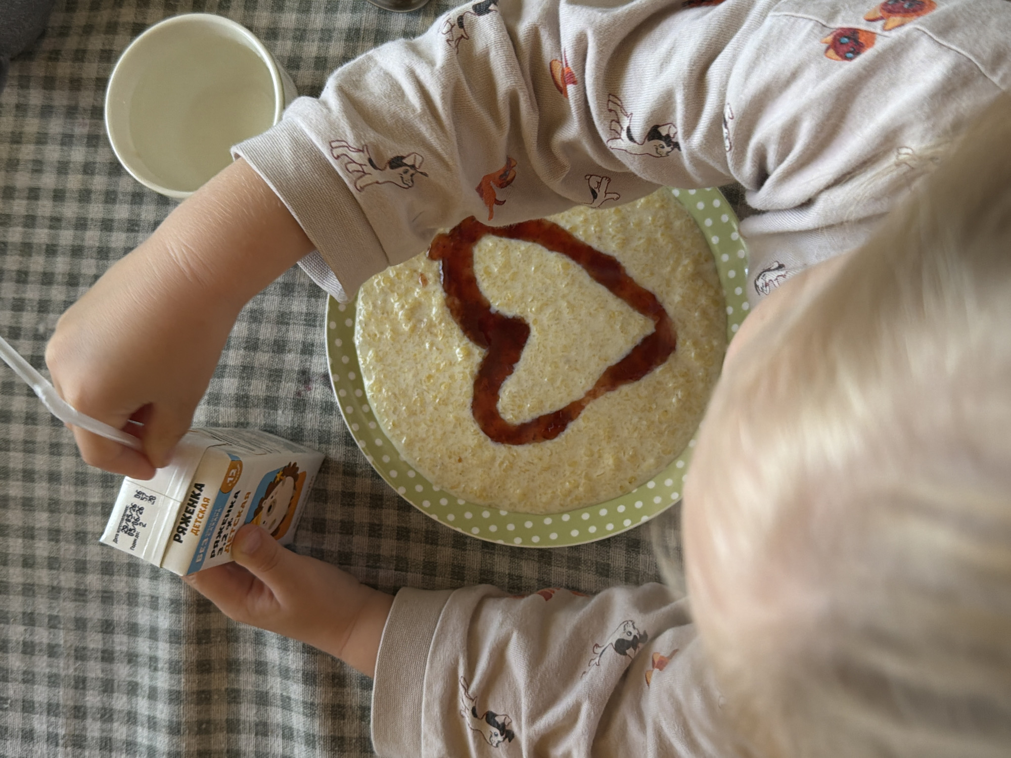 An overhead photograph depicts a child's forearms and hands engaged in a breakfast activity, positioned on a light and dark checkered tablecloth. The child wears light-colored, long-sleeved pajamas patterned with small, stylized cartoon animals, including dogs and cats in black, white, and orange-brown hues. In the child's left hand, a small, white carton of "РЯЖЕНКА ДЕТСКАЯ" (Children's Ryazhenka), a 3.2% fat fermented milk product, is held; the carton displays Russian text and a cartoon lion illustration. The child's right hand is in the process of inserting a white plastic straw into the carton's top flap. Directly centered in the frame, a round, light-green plate with white polka dots contains a yellowish, granular porridge. A heart shape has been drawn onto the surface of the porridge using a reddish-brown viscous liquid, likely jam or fruit syrup. To the upper right of the frame, a small, white ceramic bowl or mug is visible, appearing empty, with a partial view of a silver spoon handle adjacent to it. The Russian text on the carton indicates the setting is within a Russian-speaking country. The specific location of Khimki, Russia, cannot be confirmed from the visual evidence within the image.