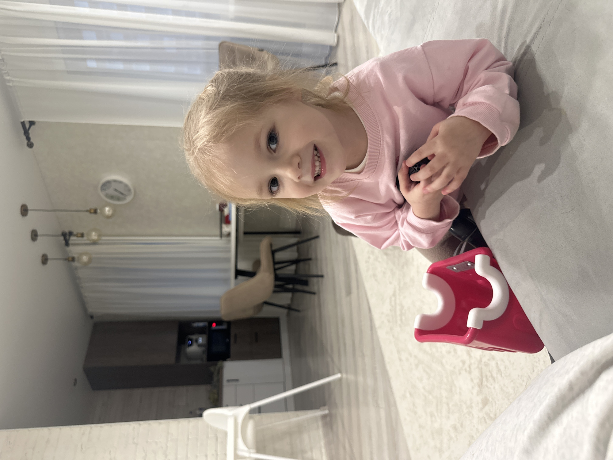A child, approximately 3-4 years old with blonde hair and blue eyes, is seated on a gray sofa. She is wearing a light pink sweatshirt. She is holding a dark object in her hands. A pink and white child's toy is to her side. The room includes sheer curtains, a dining table with chairs, and kitchen cabinetry. The room is in Chaikovsky, Russia.