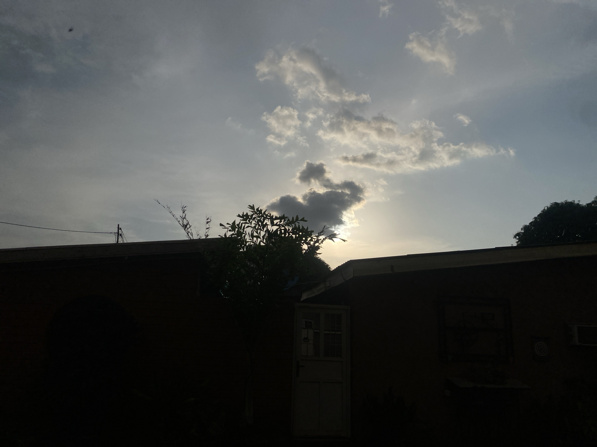 The image depicts a residential backyard in Barden Yana, Nigeria, during what appears to be dusk. A portion of a house, silhouetted against a cloudy sky, occupies the lower half of the frame. The house has a flat roof and a doorway with a multi-paned window is visible on the right side. A tree with dense foliage partially obscures the view of the house. The sky is a mix of grey and blue, with scattered clouds illuminated by the setting sun. A small, dark object, possibly a bird or an insect, is visible in the upper left quadrant of the sky. The overall impression is one of a quiet, domestic scene at the end of the day.