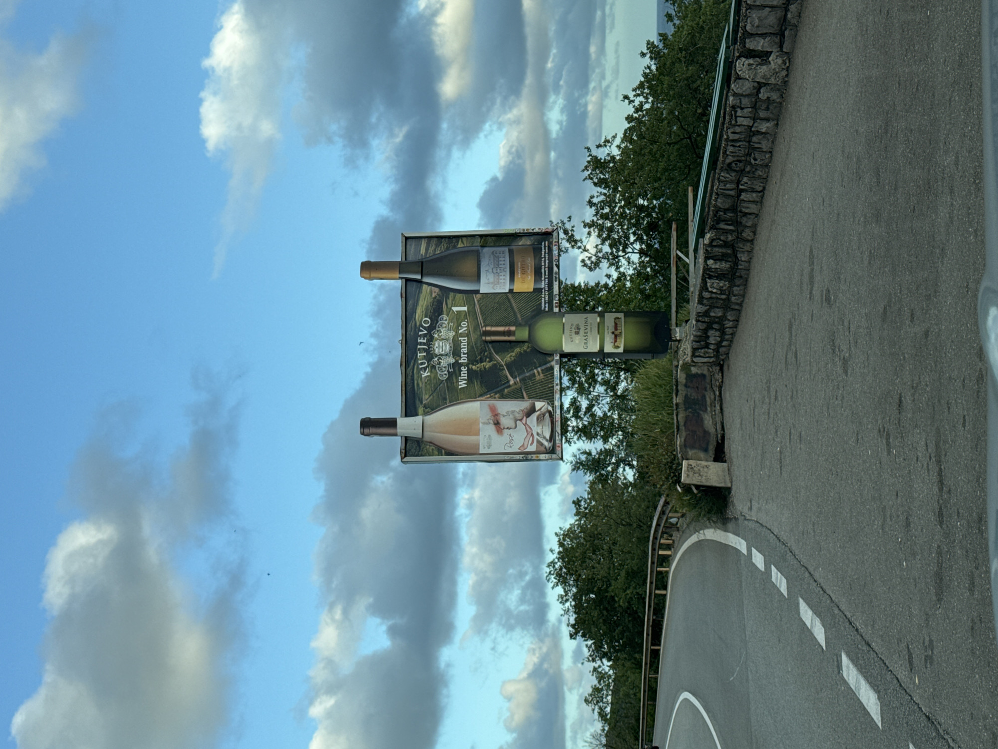 The image depicts an outdoor scene on a curved road with a billboard in Sveti Stefan, Montenegro. The road has painted white markings, and a stone wall borders it on one side, separating it from greenery. The billboard displays images of wine bottles and is titled "KUTJEVO Wine Brand No. 1". The sky, partially visible behind the billboard, is filled with clouds and blue patches.