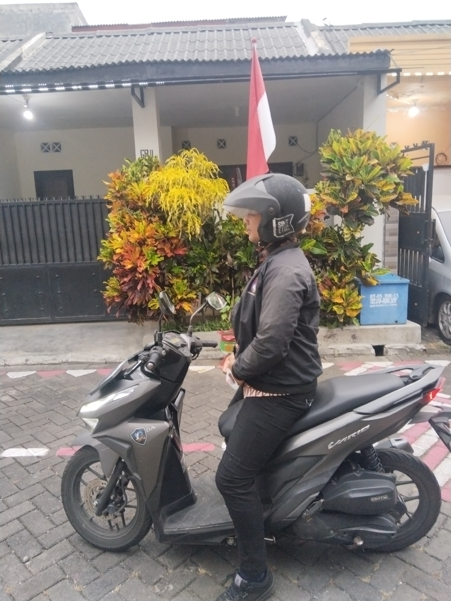 In the city of Watugede, Indonesia, a person is seated on a dark gray scooter. The individual is wearing a black helmet with a sticker that reads "SINT ETIC", a black jacket, and black pants. The scooter is parked on a paved surface made of interlocking bricks, with a white and red striped pattern visible in the foreground. Behind the scooter, a house with a gray tiled roof and beige walls is visible. In front of the house, there is a dense green and yellow bush. A red and white Indonesian flag is planted to the left of the bush. A blue recycling bin with white text is positioned to the right of the bush. The scene appears to be on a cloudy day, with diffused lighting.