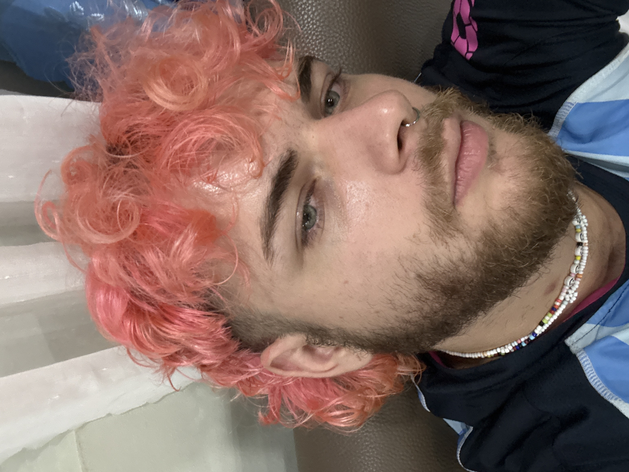 A close-up, eye-level shot features a male individual with bright pink, curly, mullet-style hair and a light brown beard. He has light-colored eyes, appearing blue or green, and a silver septum piercing. He is wearing a dark blue hooded garment with light blue and white elements, and a visible pink lining/design. Around his neck is a multi-strand beaded necklace, primarily white with colorful accents and discernible smiley face patterns. The individual looks directly towards the viewer with a neutral expression. The background indicates an indoor setting, with a brown upholstered surface, likely a couch or chair, to the left, and a light-colored, draped fabric, resembling a curtain, visible behind the individual's head and right shoulder. No specific visual cues within the image identify the location as Đà Nẵng, Vietnam.
