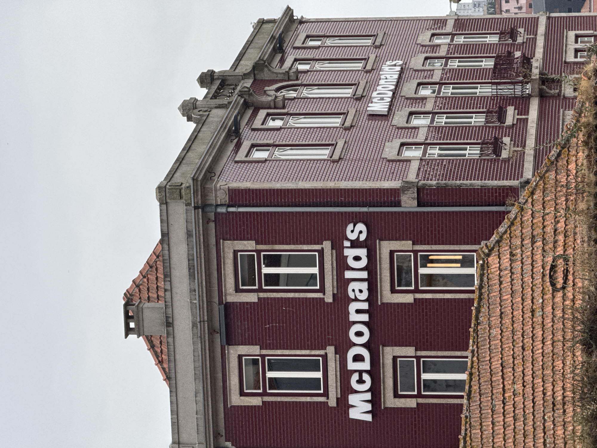 The image shows the exterior of a McDonald's restaurant in Porto, Portugal. The building is a multi-story structure with a dark red brick facade and a tiled roof. The McDonald's logo, a large white "M", is prominently displayed vertically on the building's side, spanning multiple floors. Several windows with white frames are visible on each level, some with what appear to be railings or decorative elements on the exterior. The architecture of the building suggests it might be an older, repurposed structure adapted for the restaurant. The sky is overcast, indicating a cloudy day. The image is taken from a slightly elevated angle, looking up at the building. The overall scene is urban, focusing on the commercial establishment. No people are visible in the image. The visible text is "McDonald's".