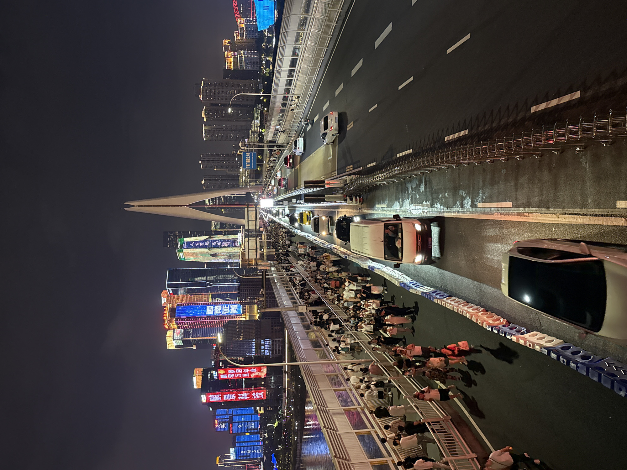 This image captures a nighttime scene on a bustling bridge, likely the Hongyadong Bridge in Chongqing, Yuzhong District, China, given the illuminated skyscrapers in the background. A wide pedestrian walkway on the left is densely populated with people walking in both directions, many appearing to be tourists, with some holding bags and taking photos. To the right of the walkway, a multi-lane road carries traffic, with several cars and a van visible. The road is wet, suggesting recent rain, and illuminated by streetlights and the vibrant lights of the surrounding city. The architecture of the city, characterized by towering modern buildings adorned with colorful neon signs and lights, dominates the skyline. A distinctive white, arching structure, part of the bridge's design, rises prominently against the night sky. The overall atmosphere is one of urban dynamism and lively activity.