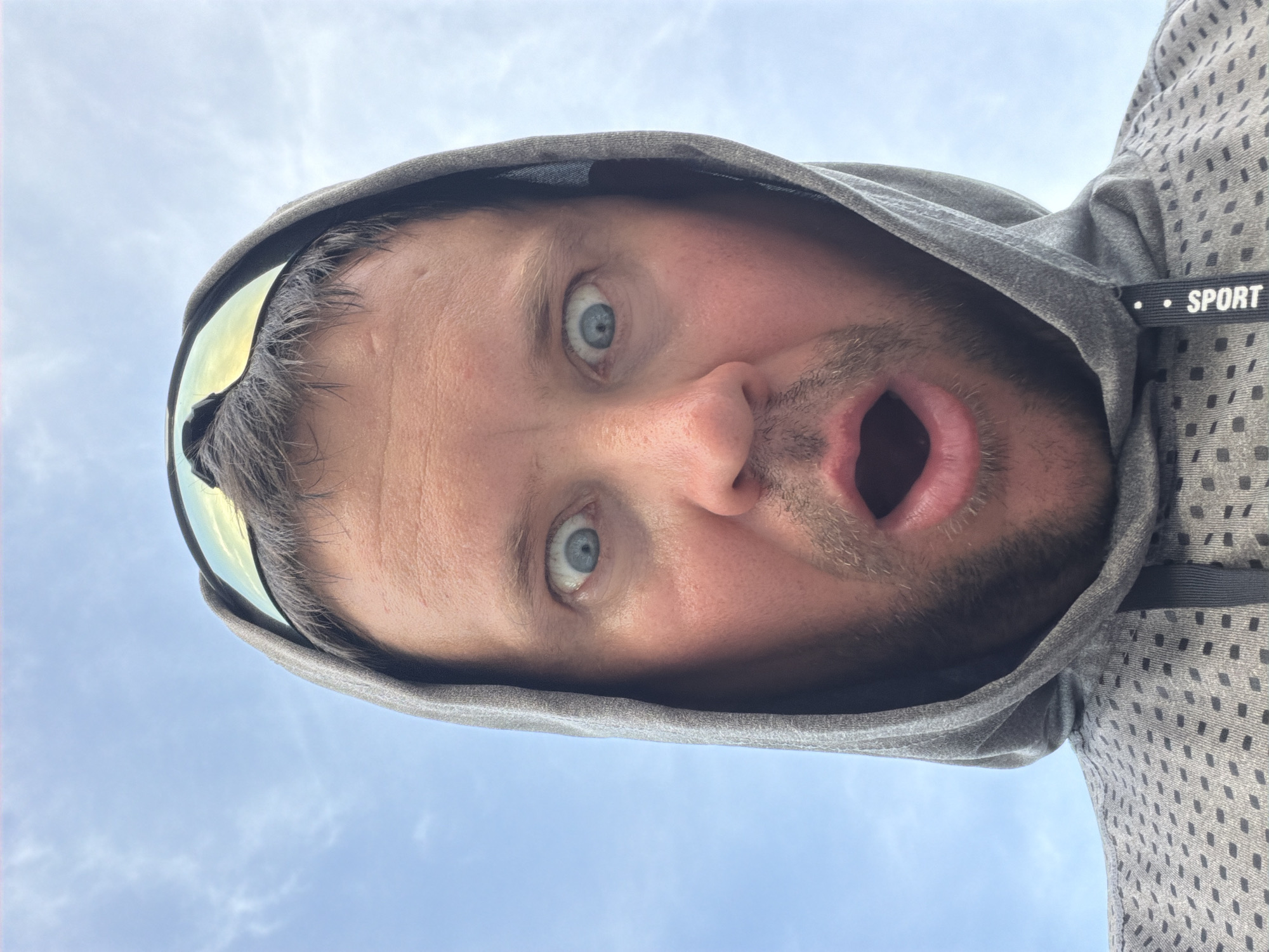 A man with a surprised expression, wearing a gray hooded sweatshirt and sunglasses perched on his head, is shown from a low angle against a cloudy blue sky. The man's eyes are wide open, and his mouth is agape. The sweatshirt has a patterned texture and a tag that reads "SPORT". The image appears to be a selfie, taken outdoors. The location is Baan Mae Nam, Thailand.