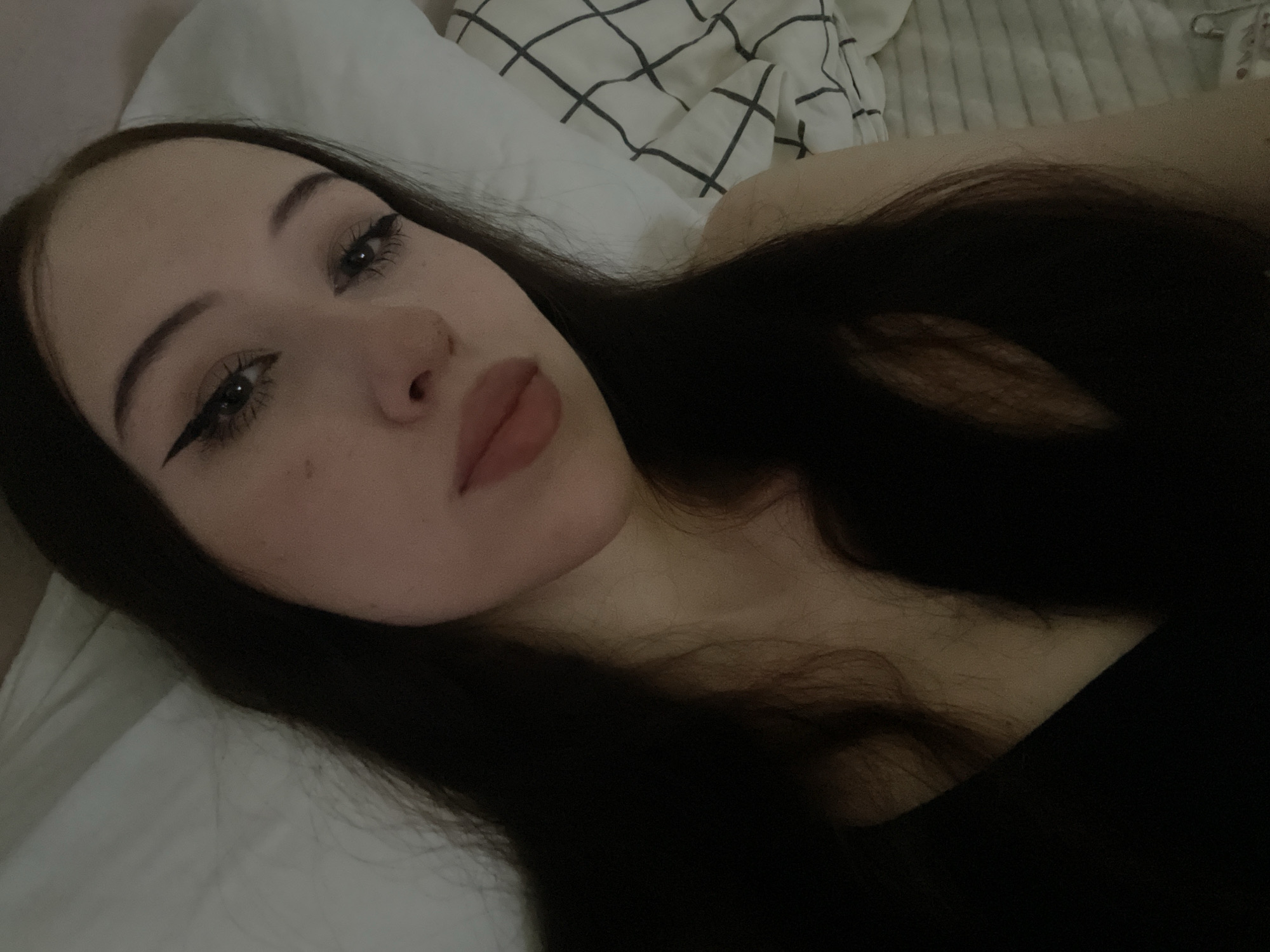 The image is a close-up, self-taken photograph of a young woman with long, dark hair, lying down. She is wearing black clothing. Her eyes are adorned with prominent winged eyeliner, and she has fair skin with subtle blush on her cheeks and a matte lipstick on her lips. Her gaze is directed towards the camera. The background consists of a white pillow and a white and black checkered fabric, suggesting an indoor setting, possibly a bedroom. No specific landmarks of Votkinsk, Russia are visible.