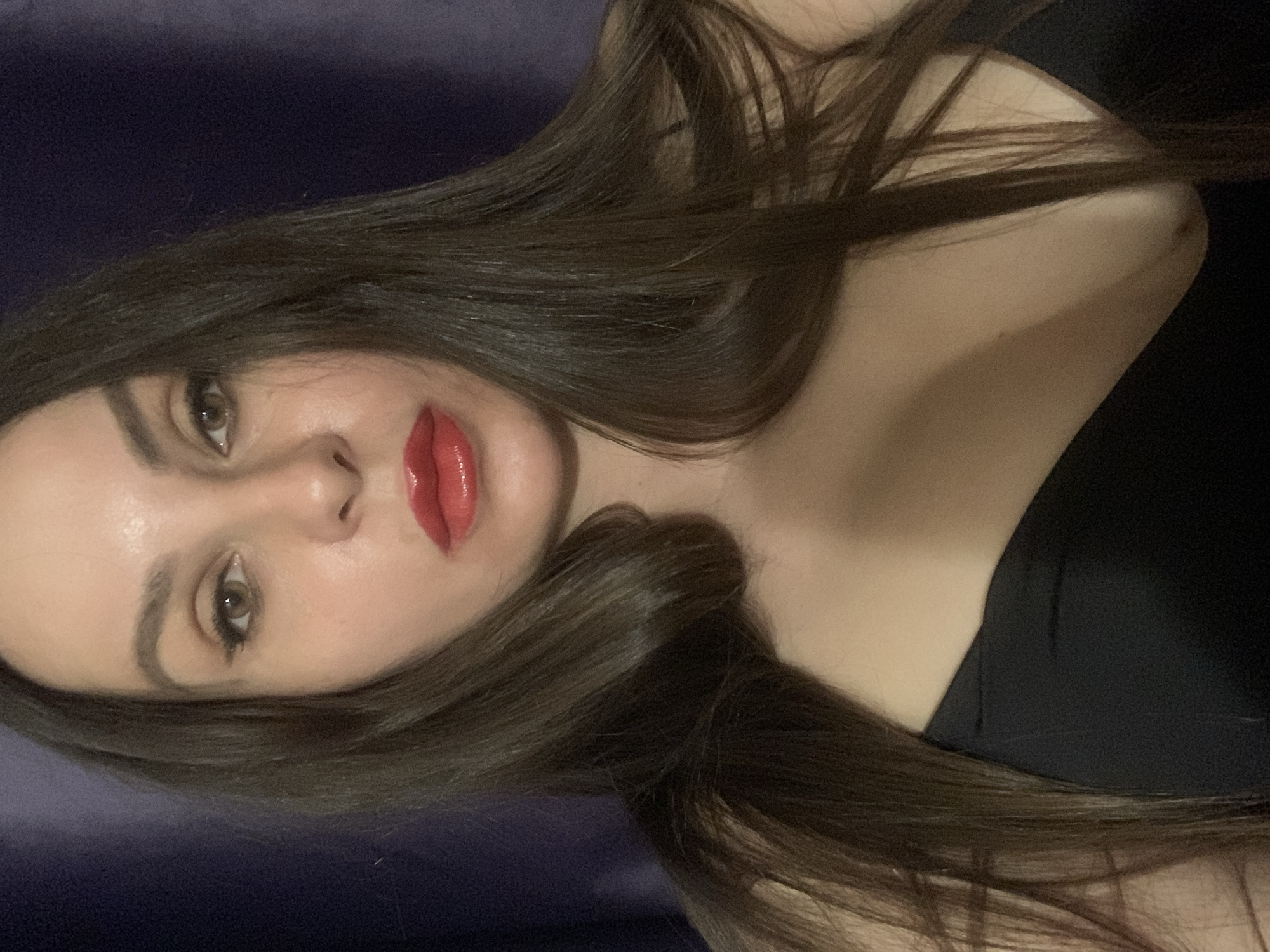 A close-up self-portrait of an adult female is depicted in the city of Unknown, Unknown. The subject has long, straight, dark brown hair parted down the middle, flowing over her shoulders and upper chest. Her face features brown eyes accentuated with dark winged eyeliner and mascara, and well-groomed dark eyebrows. She wears bright, glossy red lipstick. Her complexion appears smooth. The subject is wearing a black, strapless garment, visible across her upper chest and décolletage. The background consists of a dark, solid purple-blue textile, possibly a curtain, out of focus. The lighting is frontal and even, casting minimal shadows. The subject maintains a direct gaze with a neutral expression.