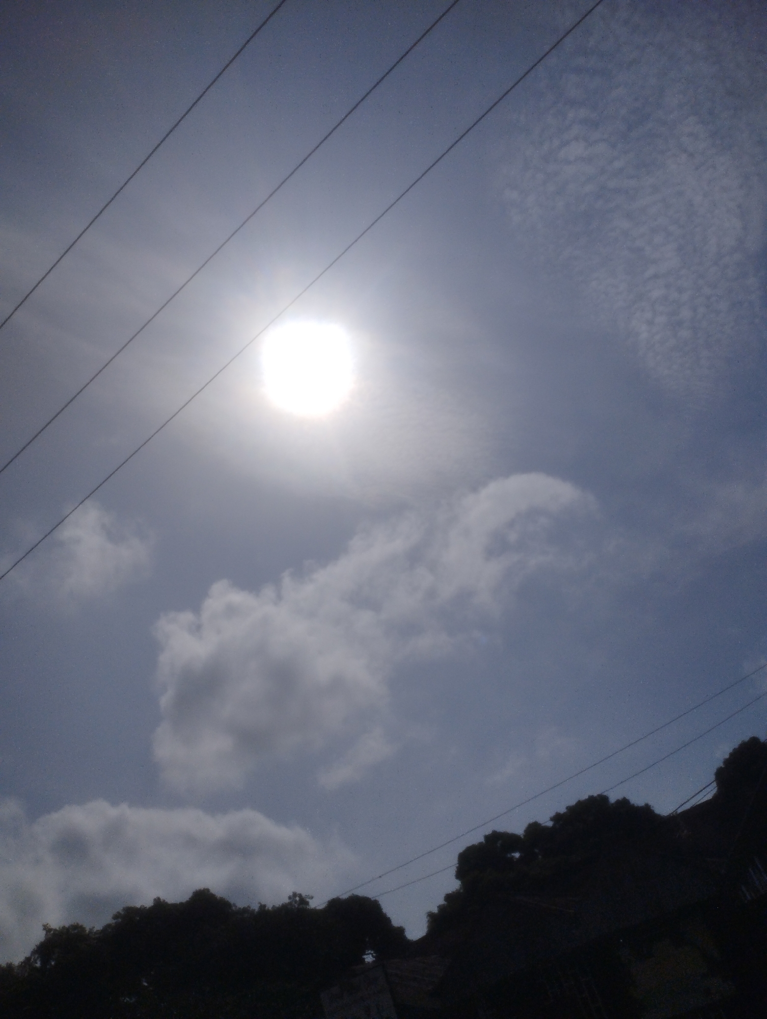 The image captures a bright, hazy sun low in the sky, partially obscured by thin clouds. Four dark power lines stretch diagonally across the frame. The lower portion of the image is dominated by the silhouetted outlines of trees and what appears to be rooftops, indicating an urban or semi-urban environment. The sky is a light blue with some scattered white clouds. There are no people visible in the image. This scene is consistent with a view from an elevated position in Jos, Nigeria, likely taken during the day when the sun is relatively low.