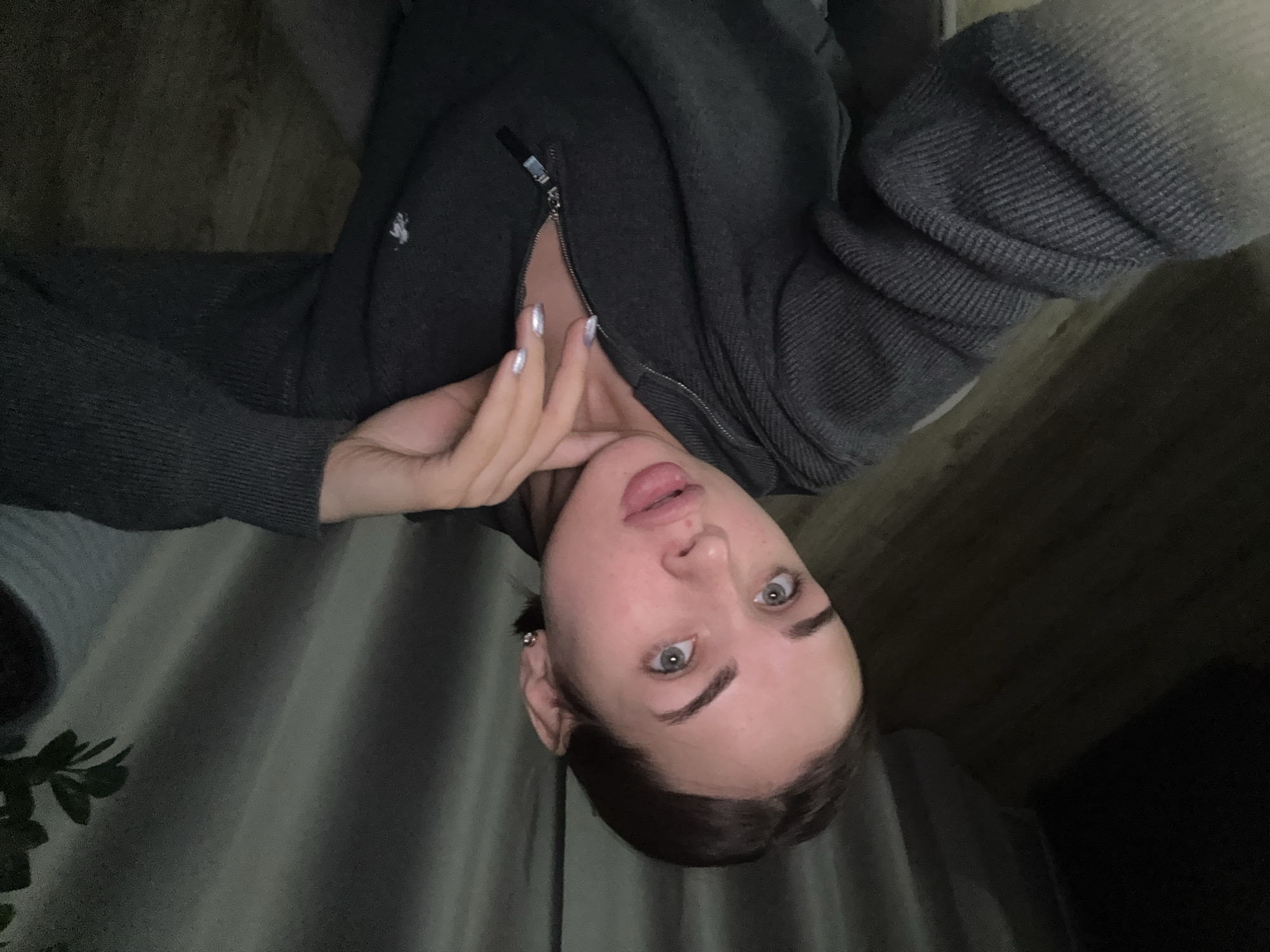 A person is shown in a selfie, tilted sideways. The person has fair skin, dark hair, and gray eyes. The person is wearing a dark gray zipped-up sweatshirt. The background appears to be a mix of curtains and dark surfaces. Based on the request for "location in the city of Лобаново, Russia" being included in the description, it can be inferred that the person is located in Лобаново, Russia.