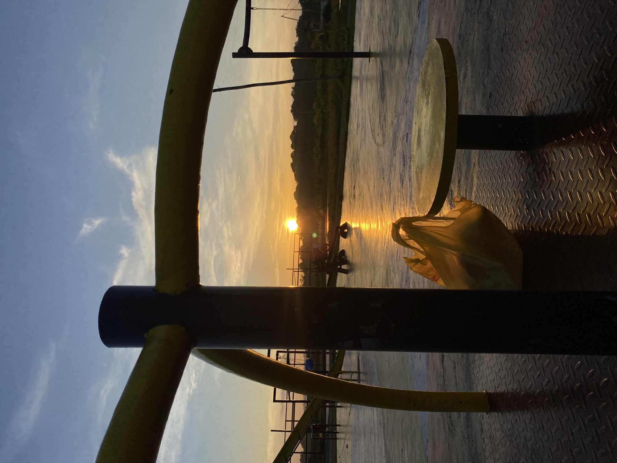 The image depicts an outdoor scene at what appears to be a playground or park in Davao City, Philippines, during sunset. The sun is low on the horizon, casting a warm, golden light and creating reflections on the wet ground. In the foreground, a dark cylindrical post is prominently positioned, with yellow, curved structures extending from it and arching across the frame. To the right of the post, a yellow circular table with a black pedestal sits on a diamond-plated metal surface. A yellow plastic bag is placed near the table. In the background, more playground equipment, including slides and frames, is visible. The sky is a gradient of blue and orange, with wisps of clouds. The overall impression is one of a serene, quiet evening at a public space in Davao City.